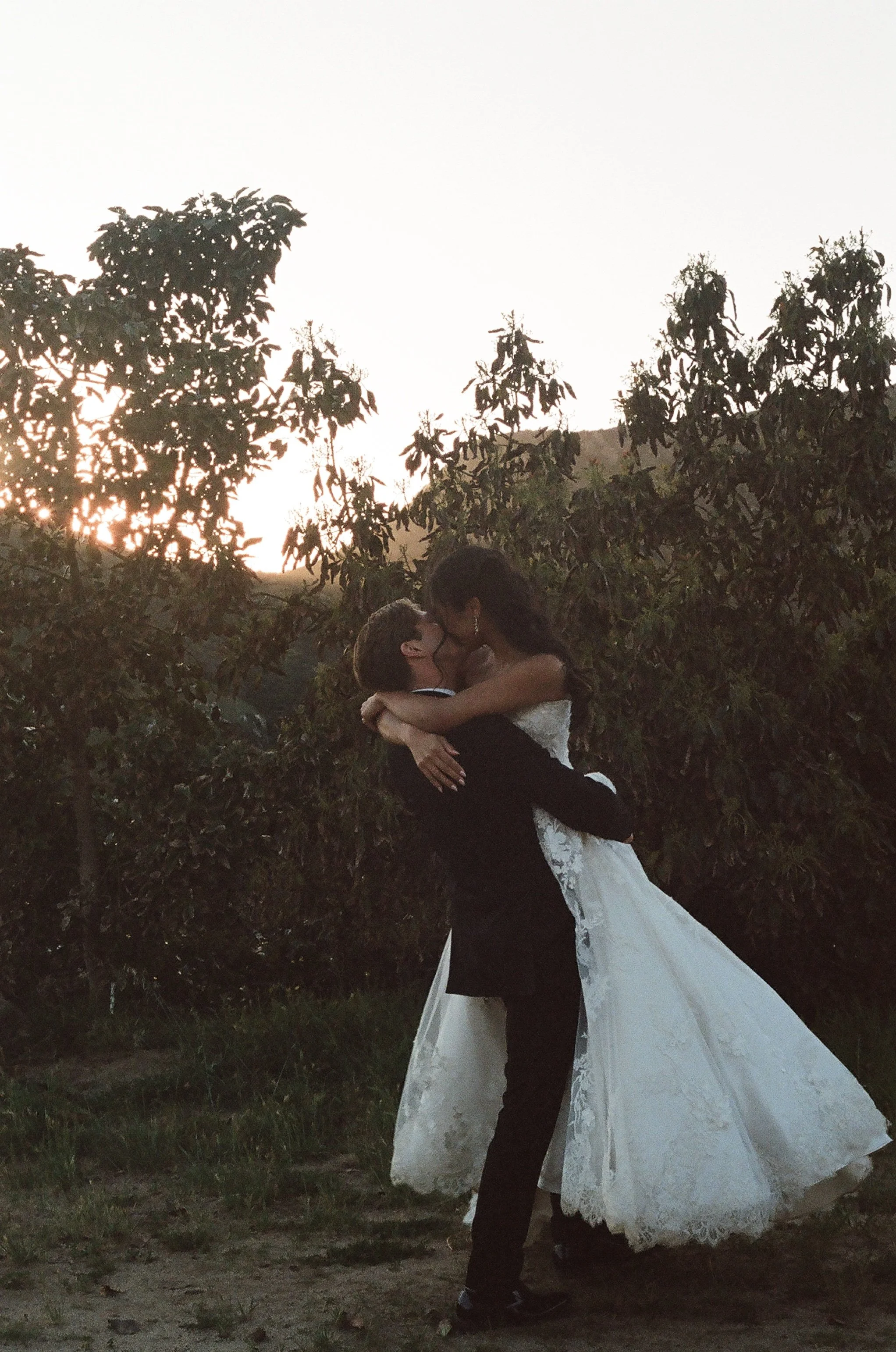A couple dressed in wedding attire, the groom lifting the bride in an outdoor setting with trees and a sunset in the background.