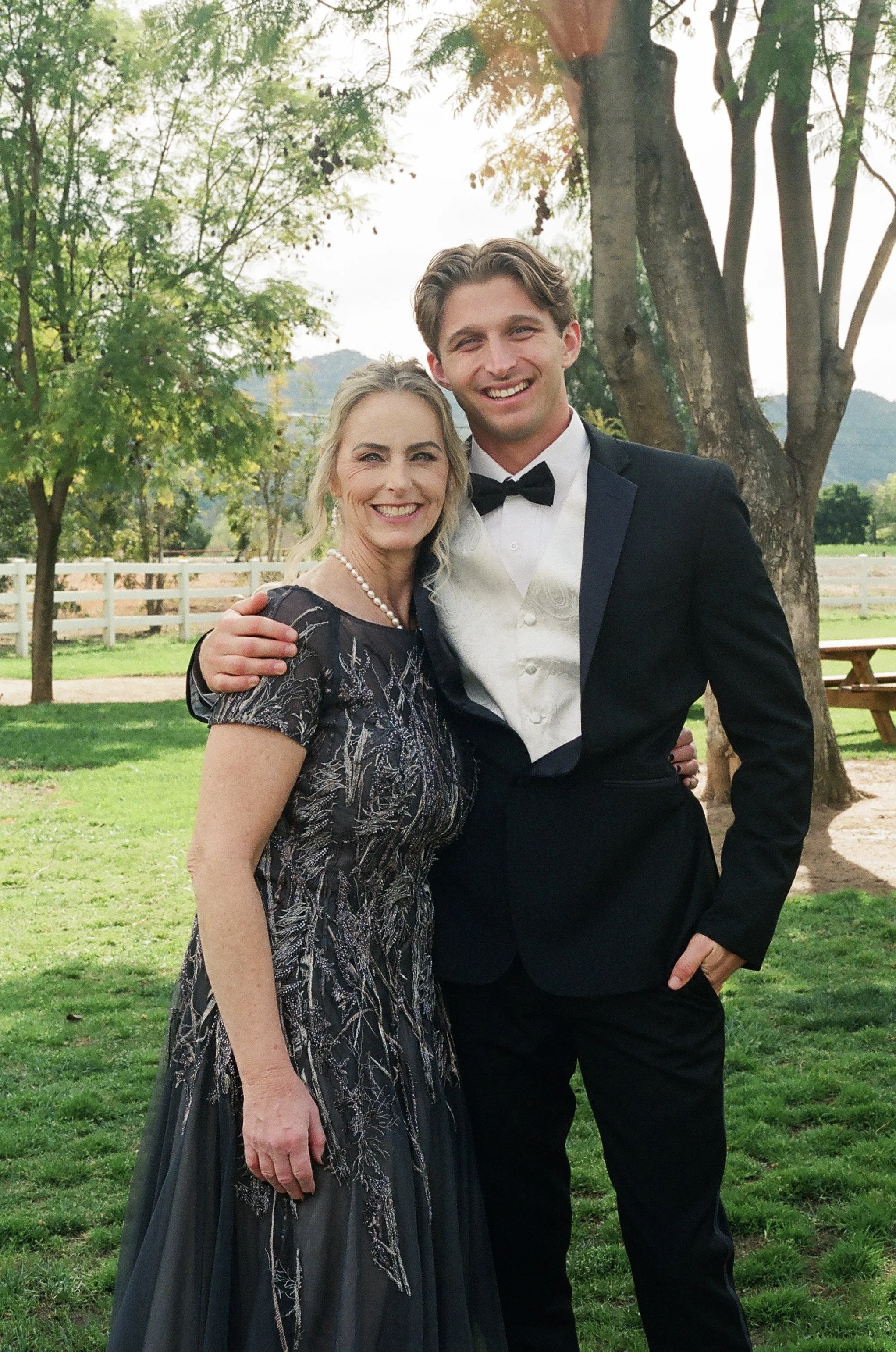 A woman and a man dressed in formal attire, outdoors, smiling and posing for a photo, with trees and a white fence in the background.