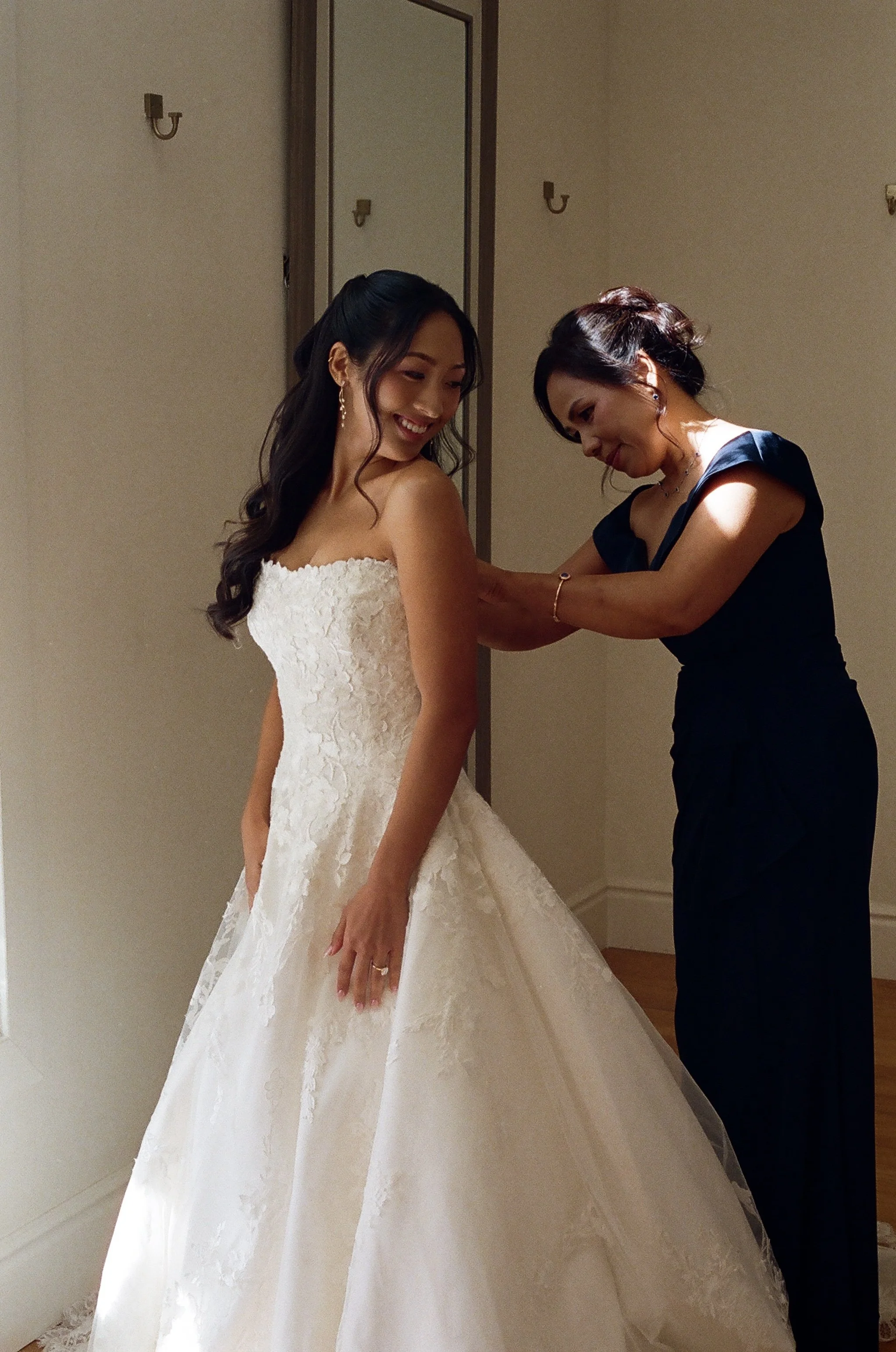 A bride in a white wedding gown standing beside a woman in a dark blue dress, who is adjusting her dress, in a room with plain walls and a wall mirror.