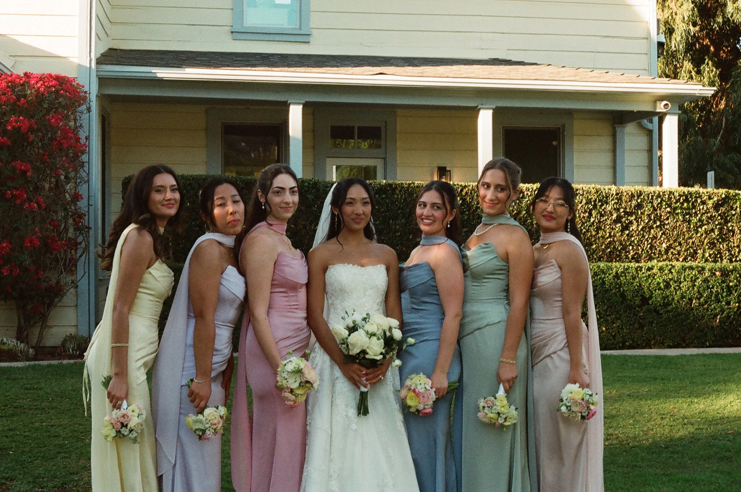A bride in a white wedding dress holding a bouquet of white roses standing among seven women dressed in pastel-colored dresses, all holding smaller bouquets, outdoors in front of a house with green siding and red flowers.