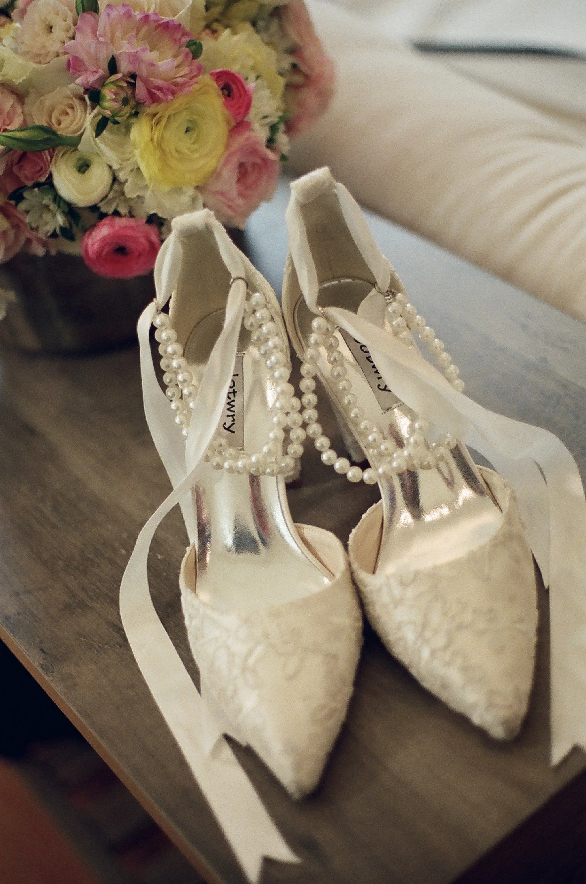 A pair of elegant champagne-colored high heels shoes with pearl decorations on the straps, placed together on a table in front of a bouquet of pink, yellow, and white flowers.