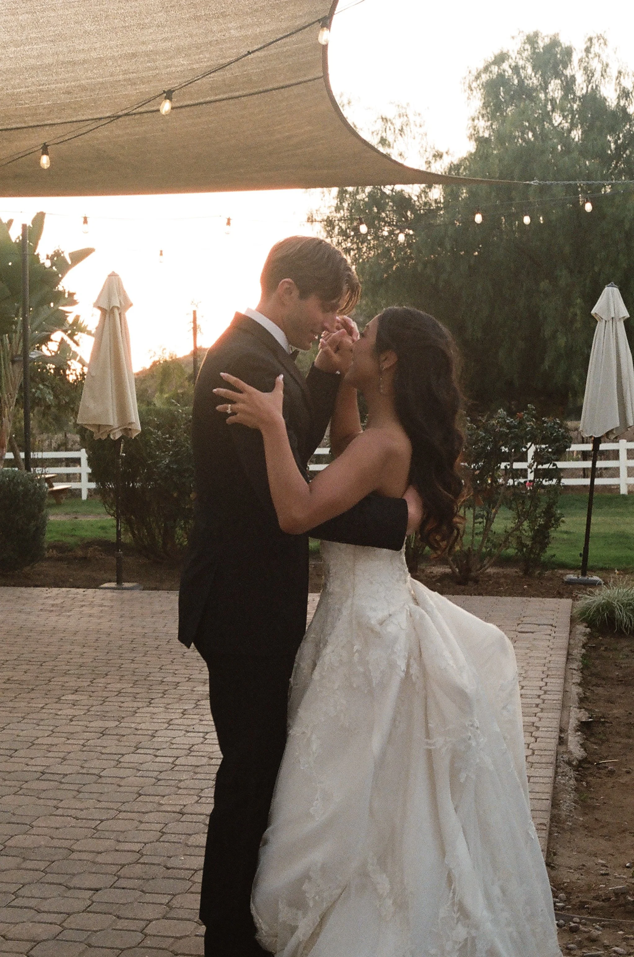 A bride and groom dancing at their wedding reception outdoors during sunset, with string lights and umbrellas in the background.