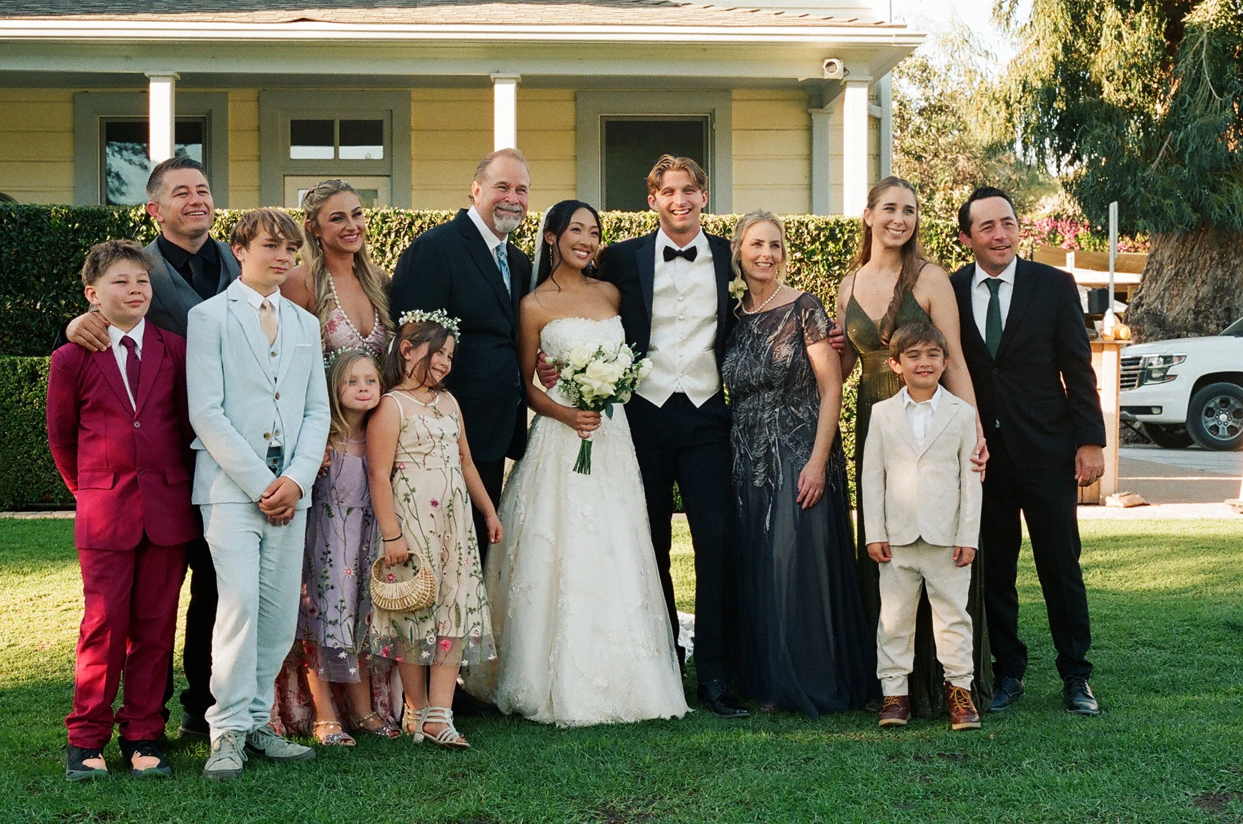 Group of people at a wedding, including the bride, groom, and wedding party, standing outdoors in front of a house with green bushes and trees.