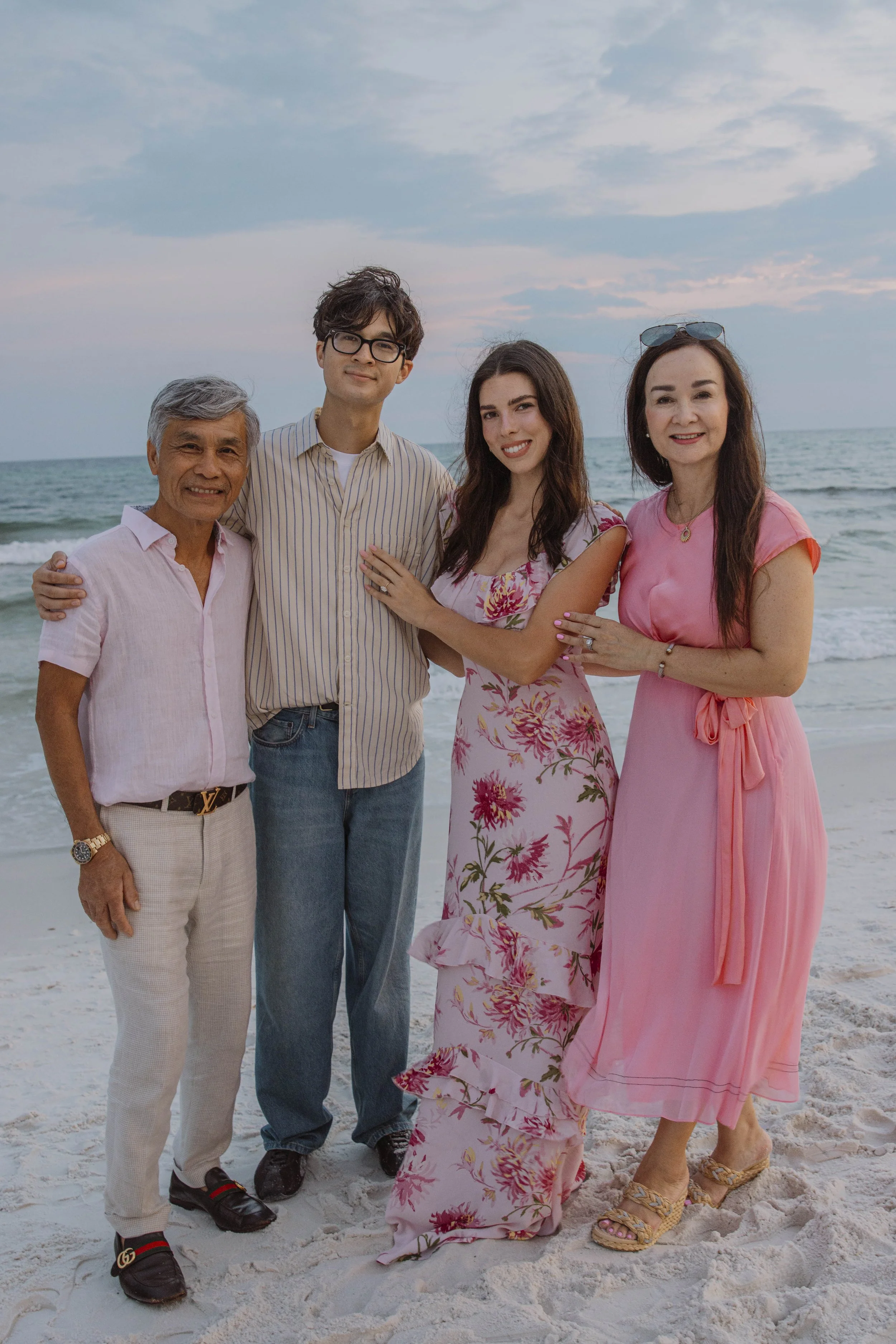 Four adults standing on a sandy beach with the ocean and sky in the background, smiling at the camera.