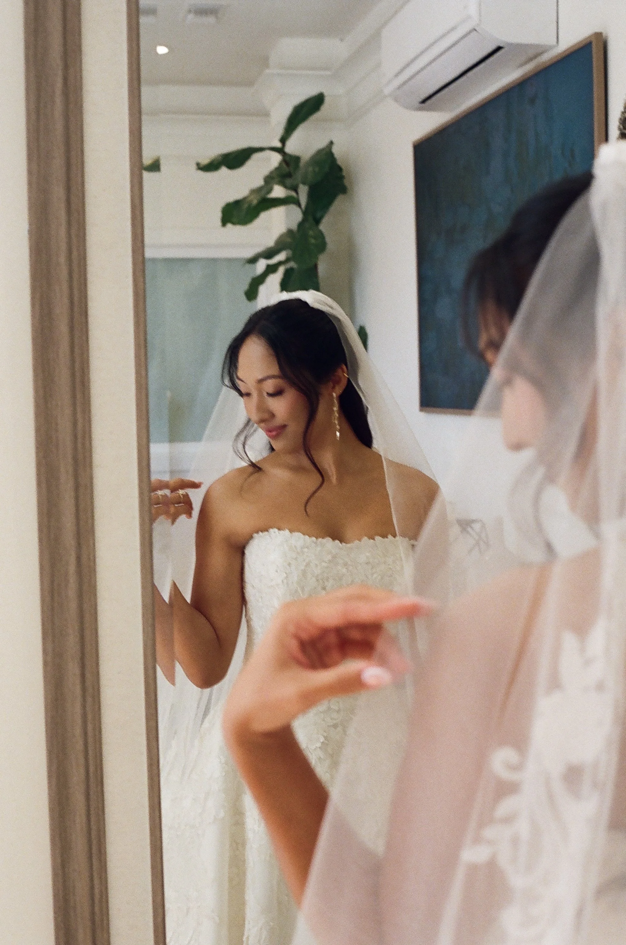 A bride in a strapless white wedding gown, looking at herself in a mirror, with a veil and jewelry, while a person in a pink shirt holds a phone in front of her.