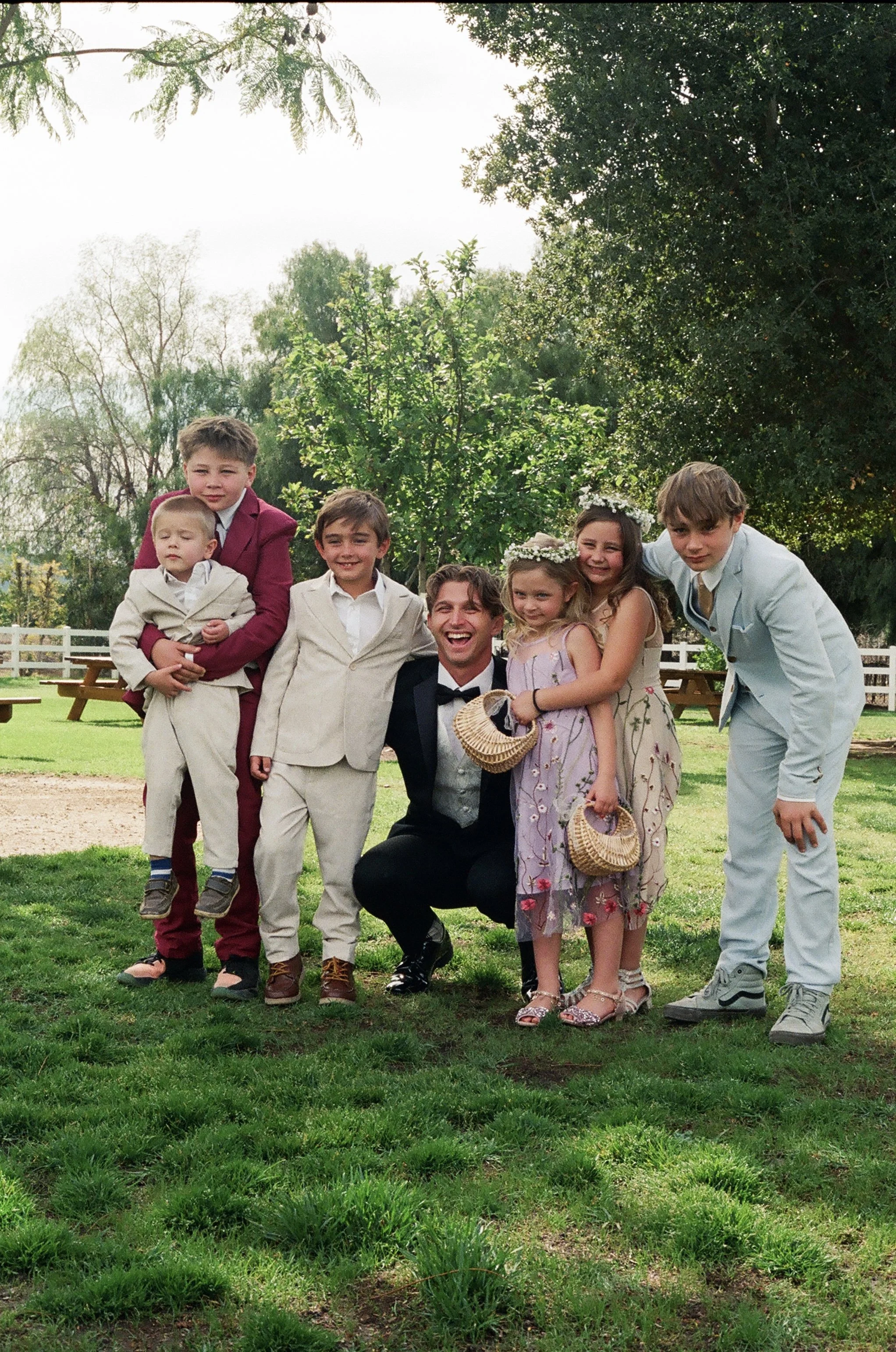 A group of children and a man posing outdoors on a grassy area with trees in the background. The children are dressed in formal or semi-formal clothing, some wearing floral or pastel dresses and suits. The man is wearing a black tuxedo with a bow tie