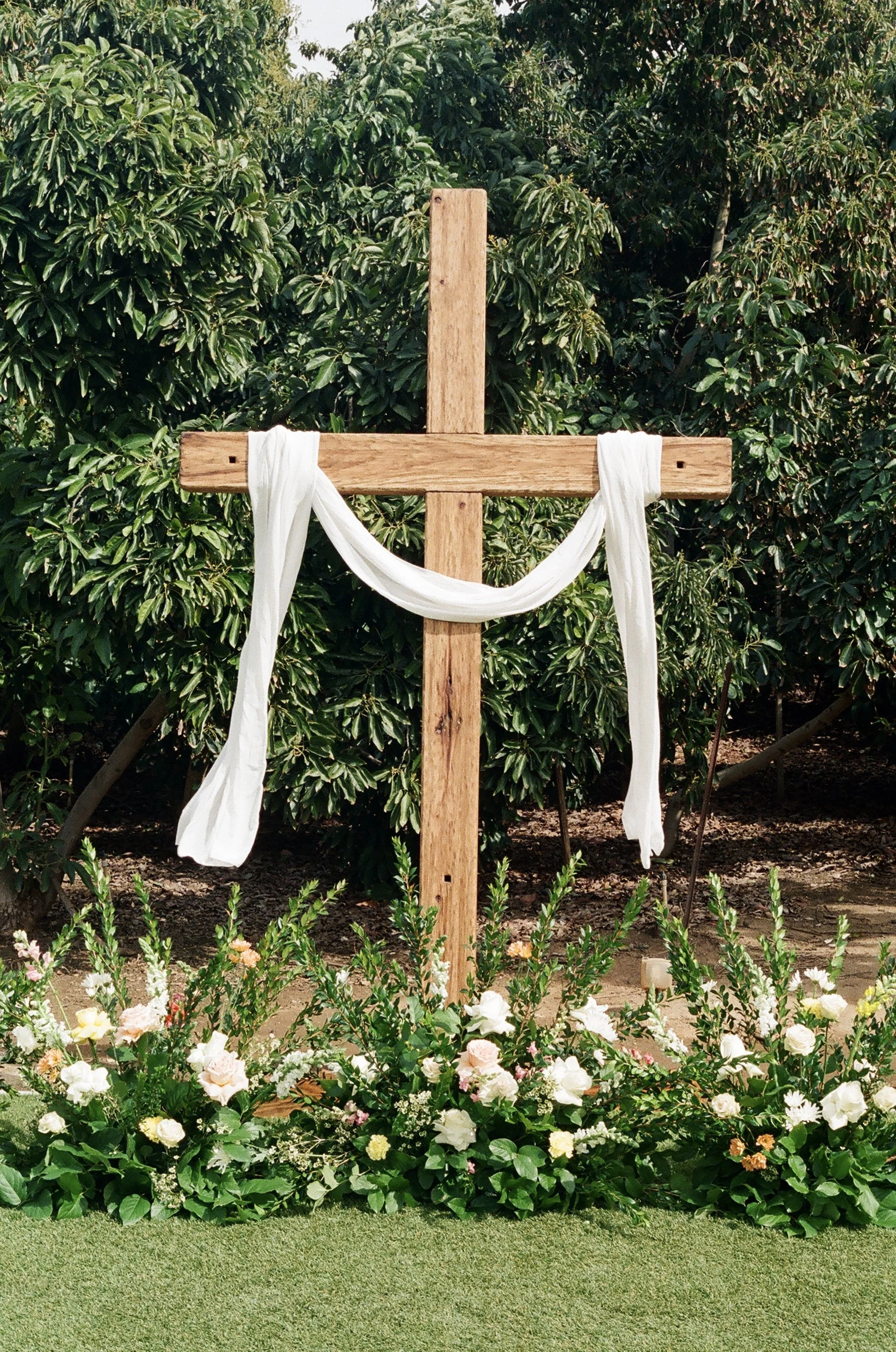 Wooden cross draped with white fabric, surrounded by flowers and greenery in an outdoor setting.