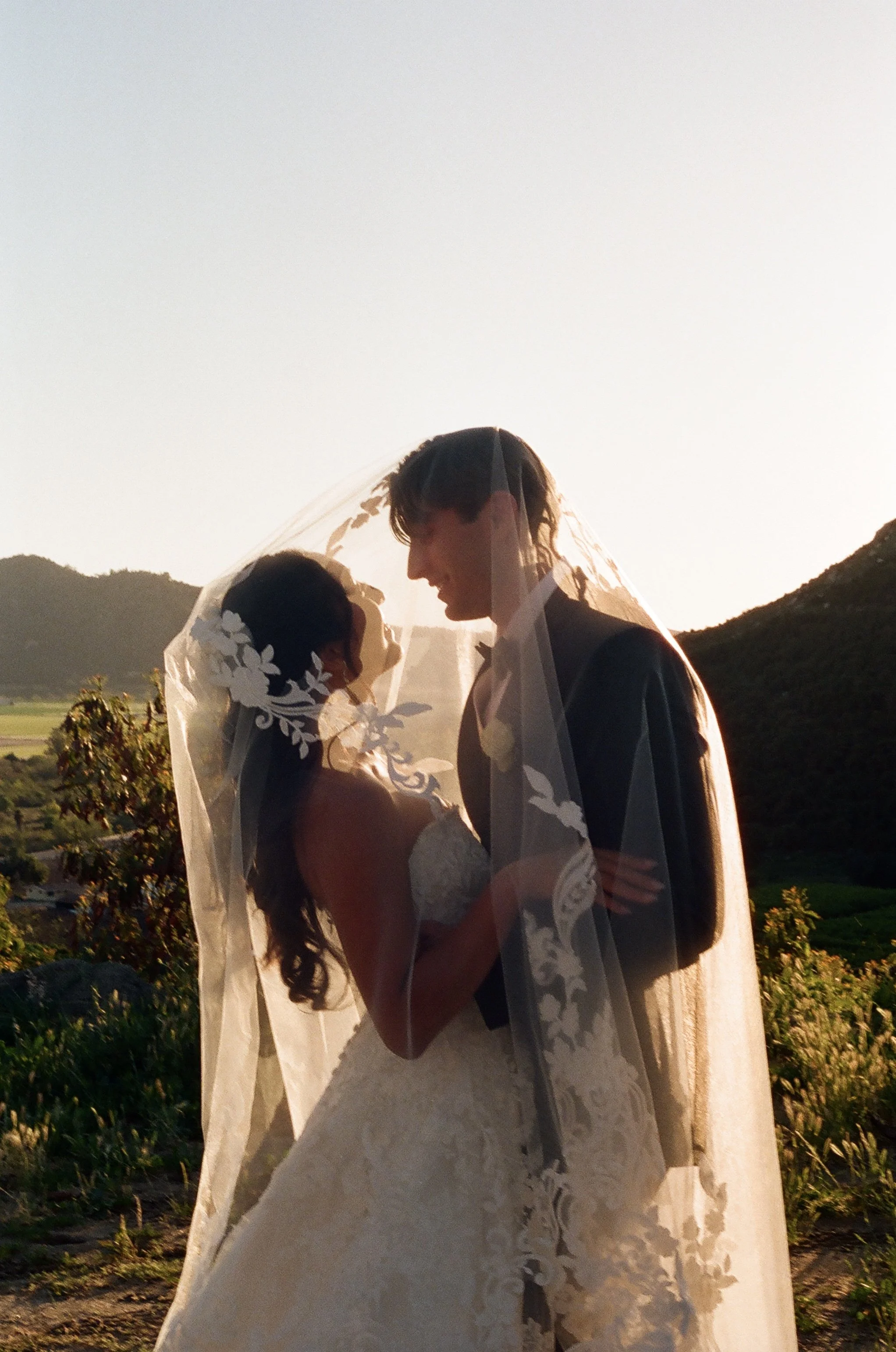A bride and groom standing close together outdoors during sunset, with mountains and greenery in the background, sharing a moment of intimacy.