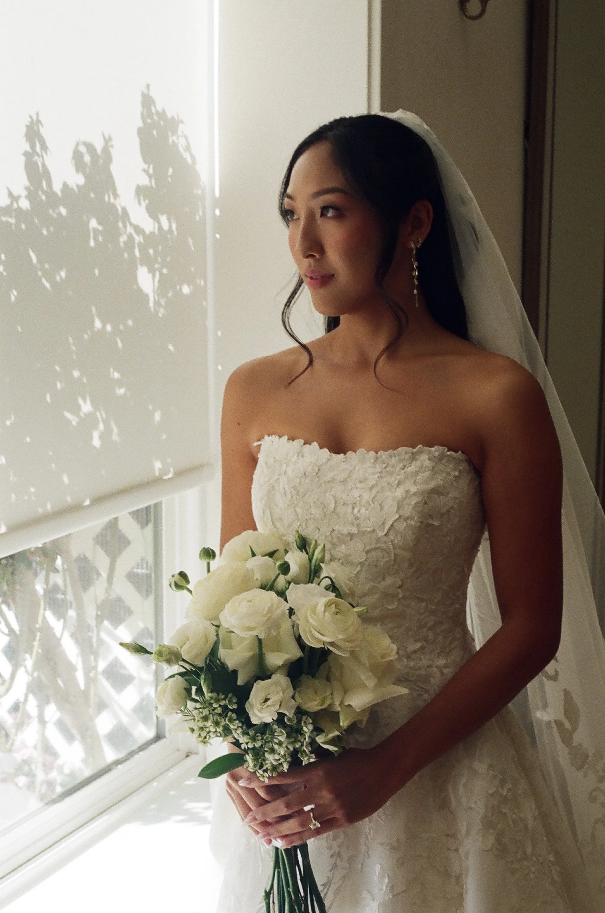 A bride in a strapless wedding dress holding a bouquet of white flowers, standing near a window with sunlight casting shadows of leaves on the wall.