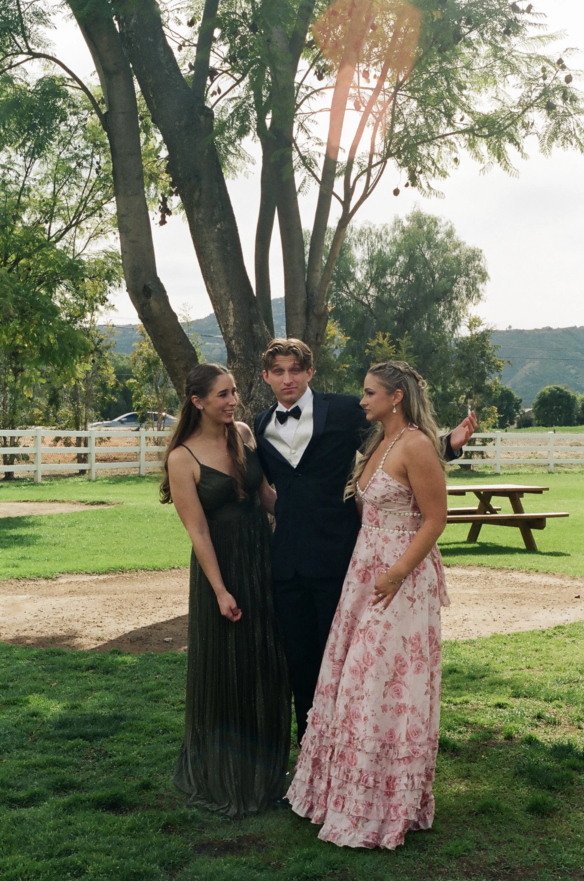 Three young people in formal attire standing outdoors near a large tree with a dirt path and green grass, with hills and a wooden fence in the background.