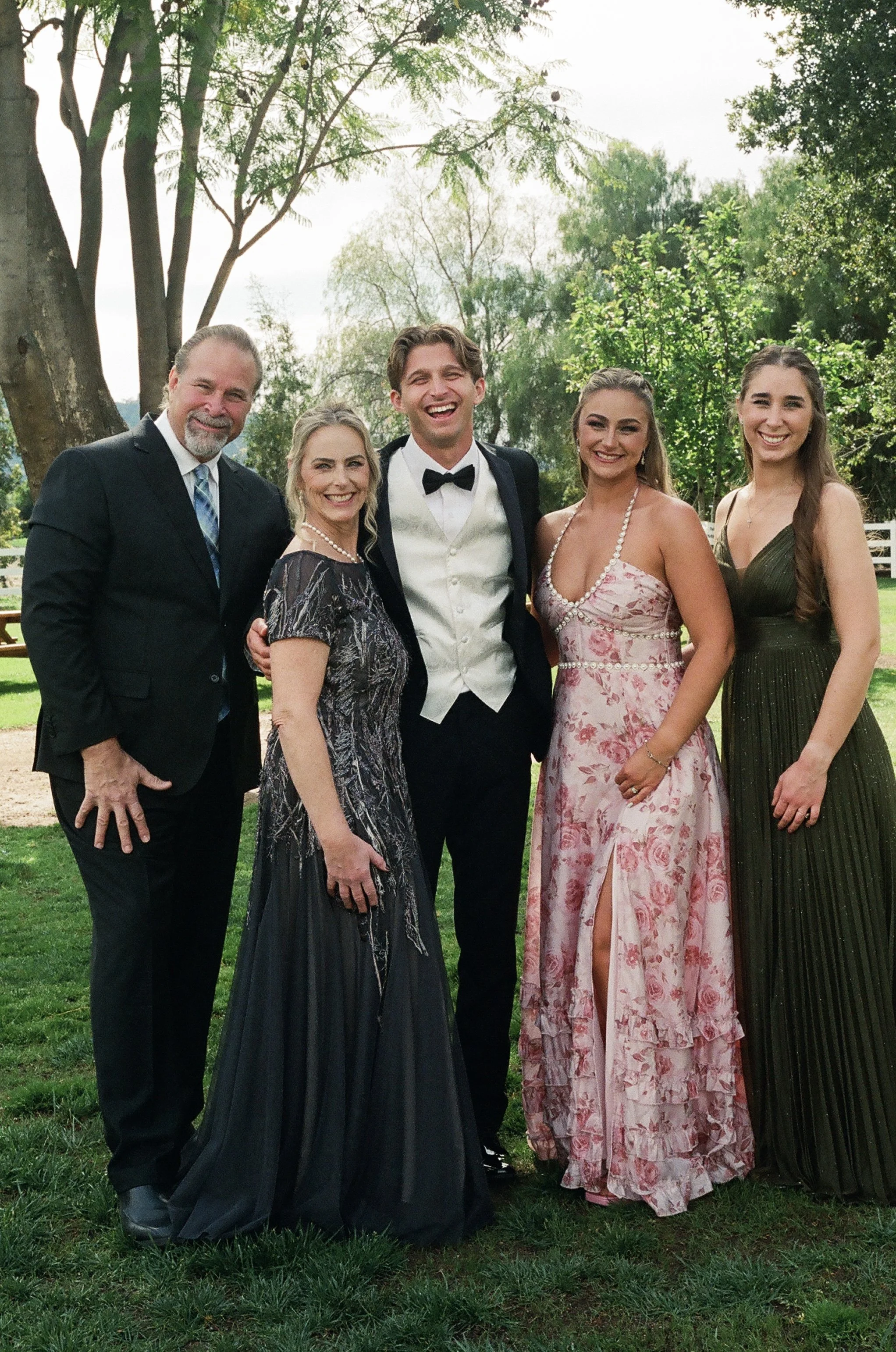 Group of five people dressed in formal attire smiling outdoors on a grassy area with trees in the background.