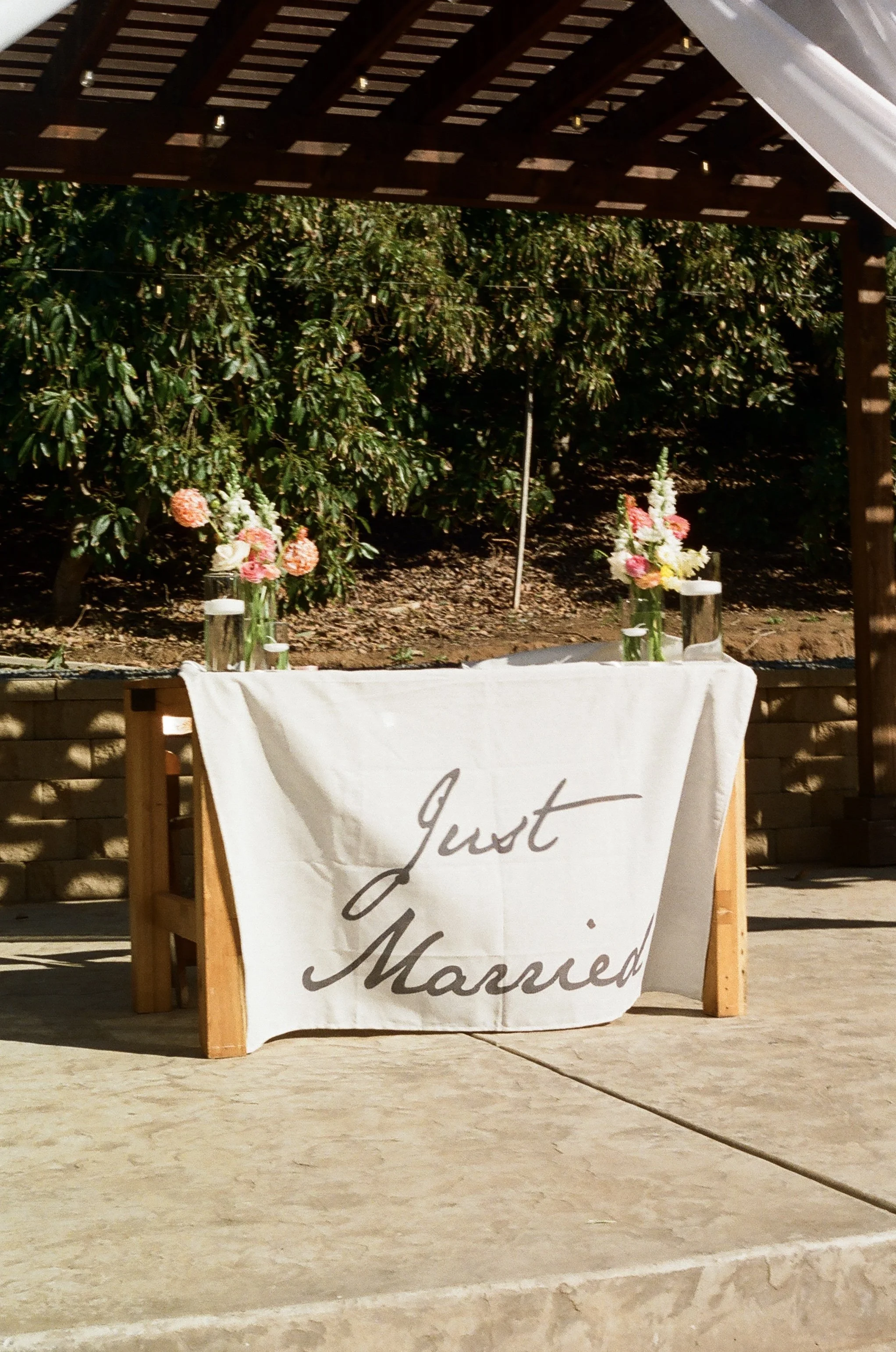 A wooden table with a white cloth that reads 'Just Married' in elegant script, surrounded by small flower arrangements in vases. The setting appears to be outdoors, with a backdrop of green bushes and a pergola.