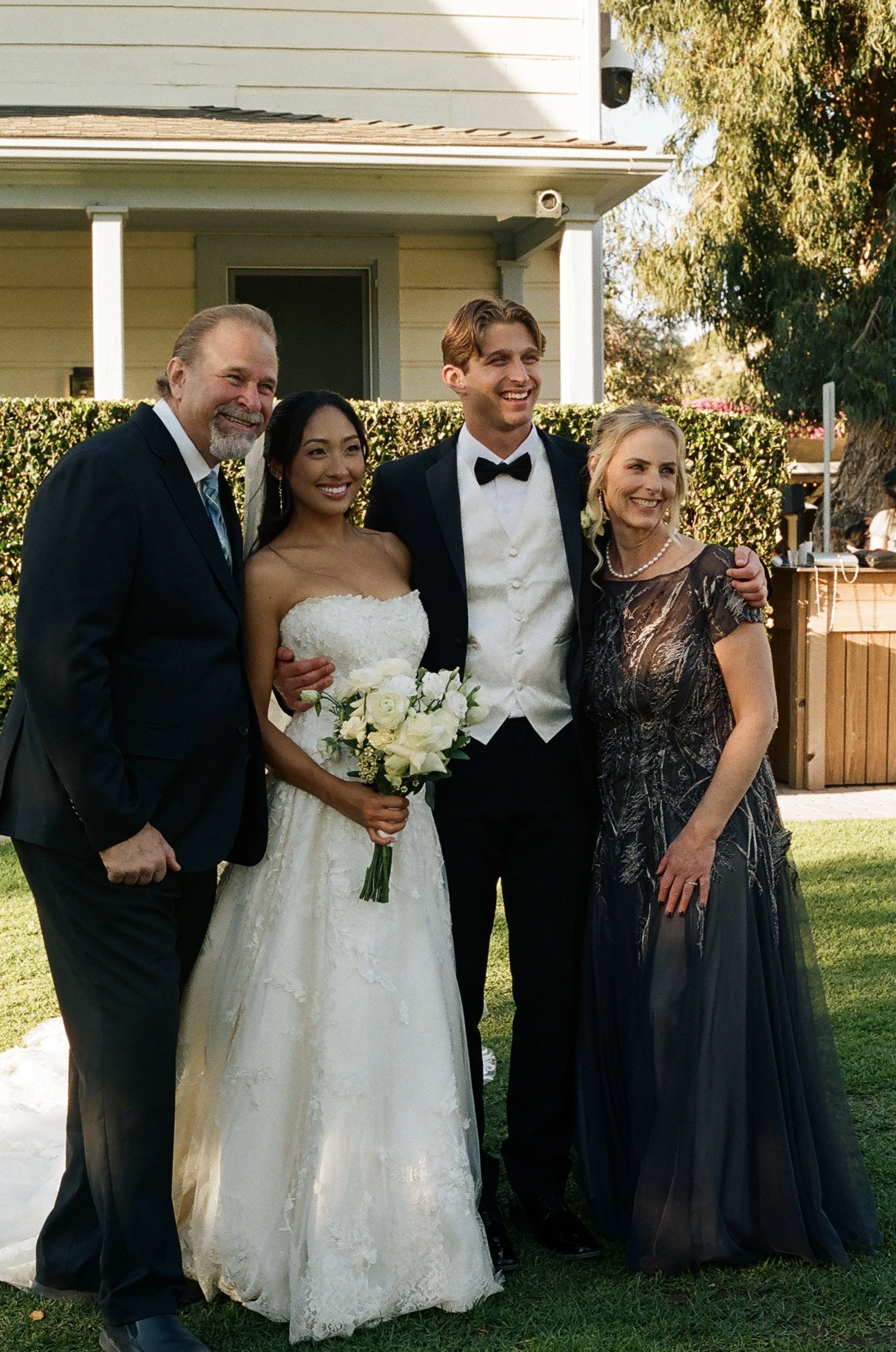 Group of five people celebrating a wedding outdoors, including a bride in a white wedding dress holding a bouquet of white roses, standing with three men and a woman in evening attire, all smiling.