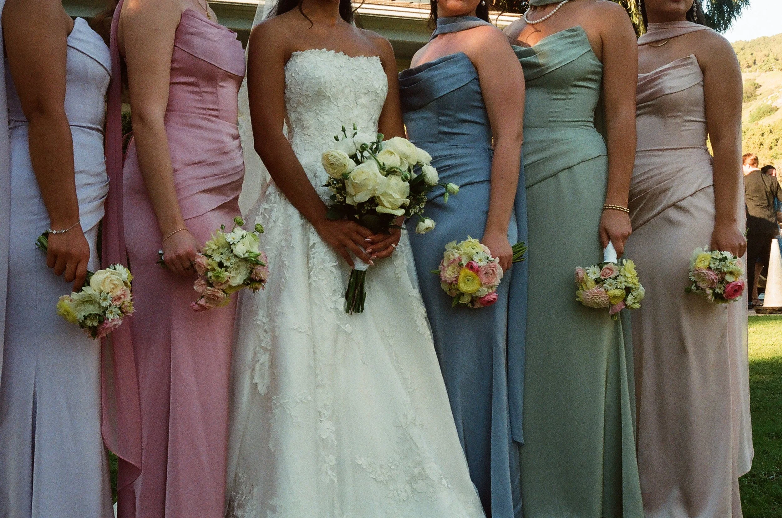 A bride in a white lace strapless wedding dress holding a bouquet of white flowers, standing among bridesmaids in pastel-colored dresses holding smaller bouquets, outdoors under a wooden structure with a natural background.