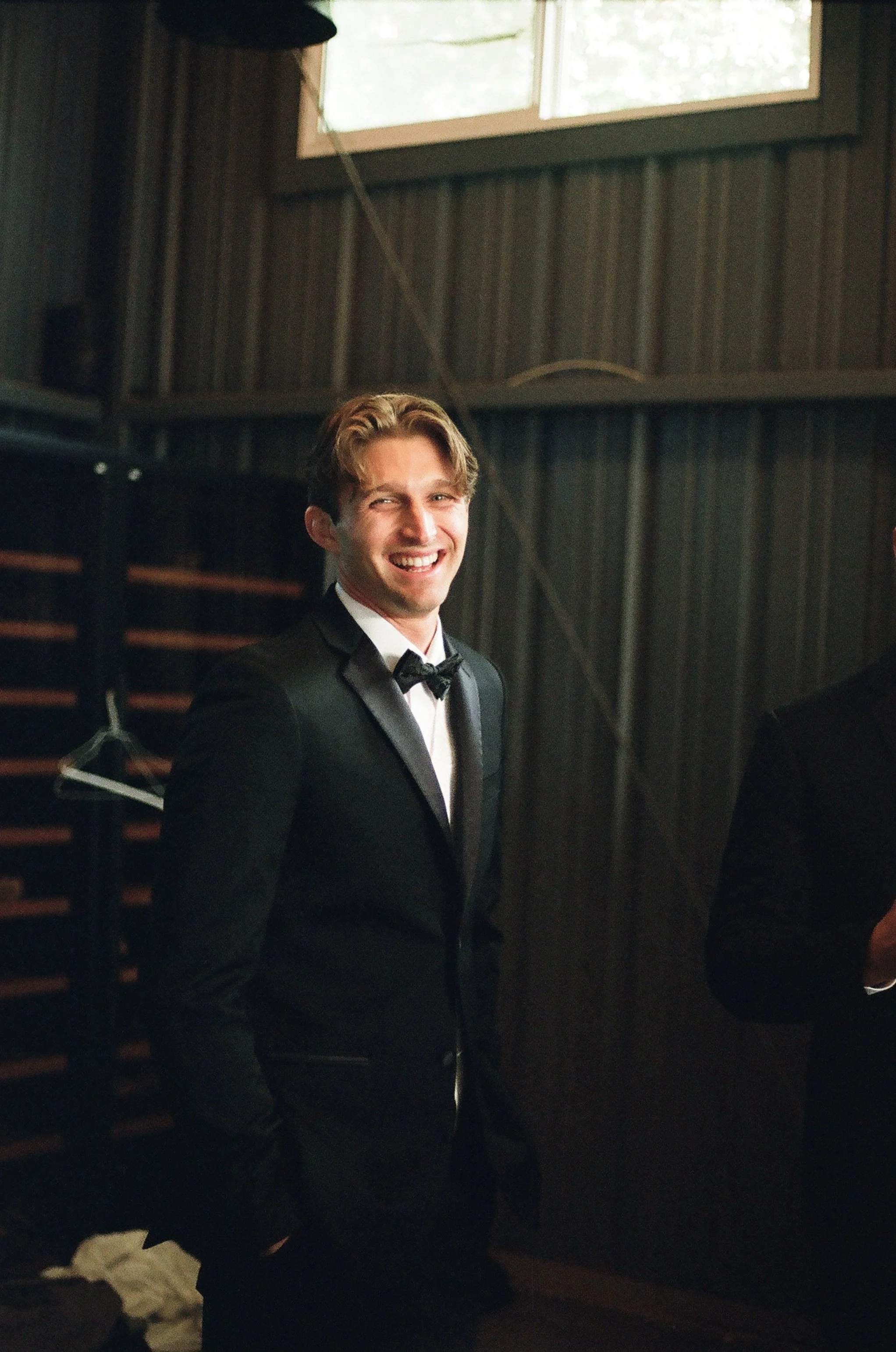 A man wearing a tuxedo and bow tie smiling inside a dimly lit room with dark wood walls and a window in the background.