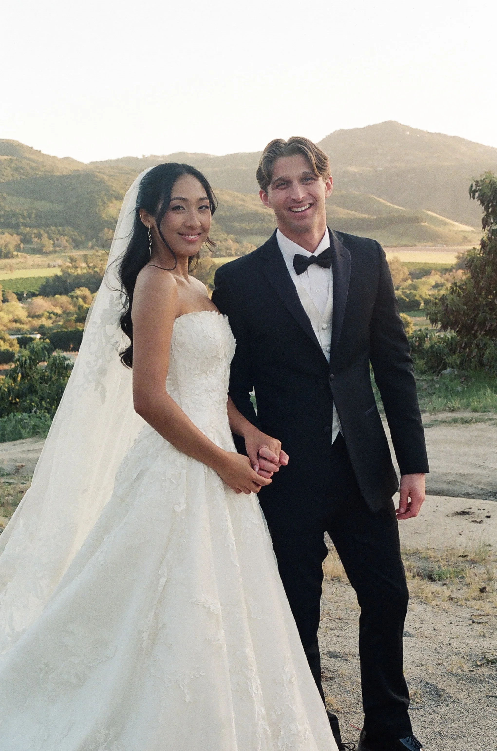 A bride and groom holding hands outdoors during sunset, with mountains and greenery in the background. The bride is wearing a white strapless wedding gown with lace details and a veil, and has long dark hair. The groom is dressed in a black tuxedo wi
