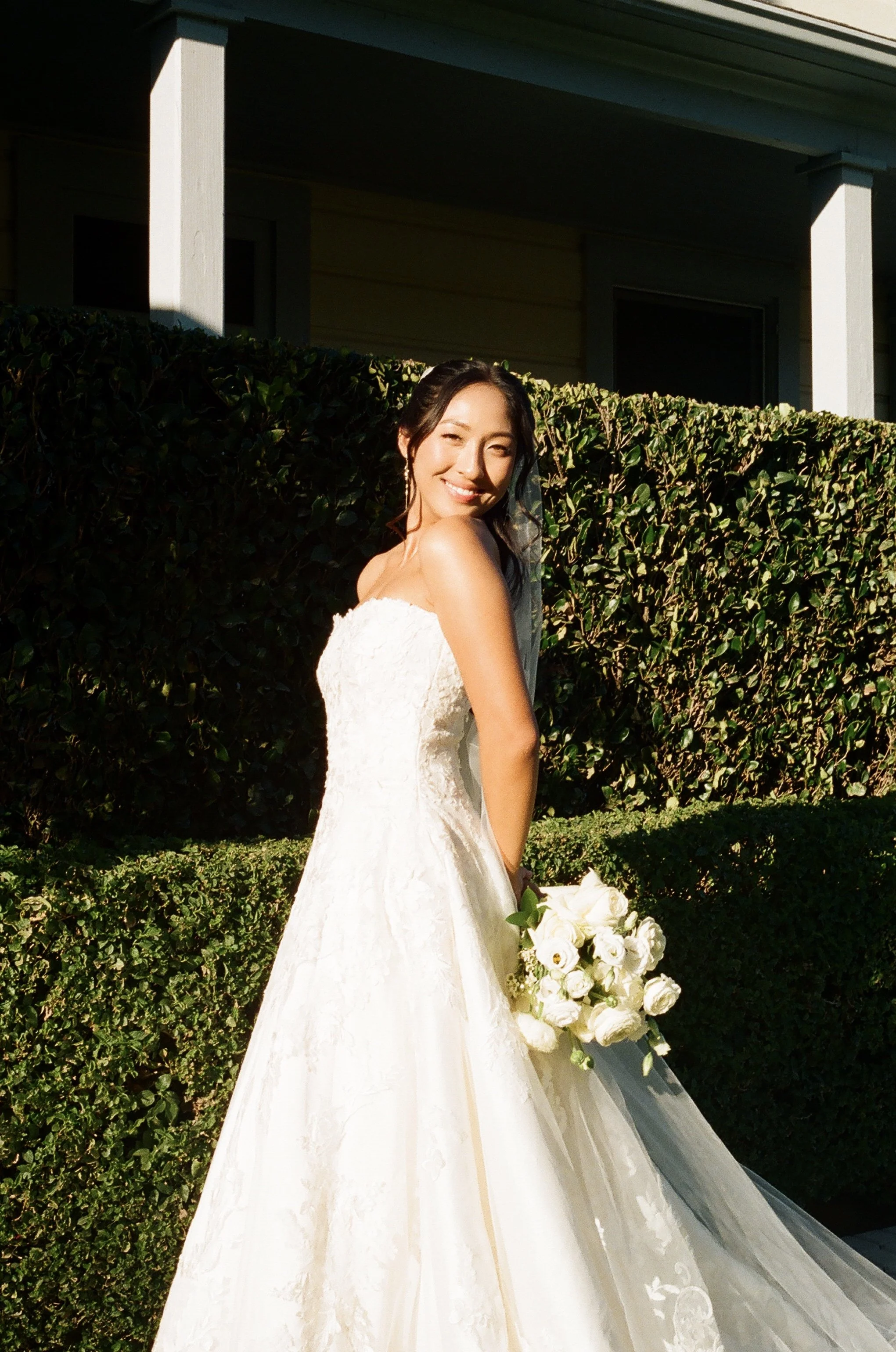 A woman in a wedding dress holding a bouquet, standing outdoors with a hedge and house in the background.
