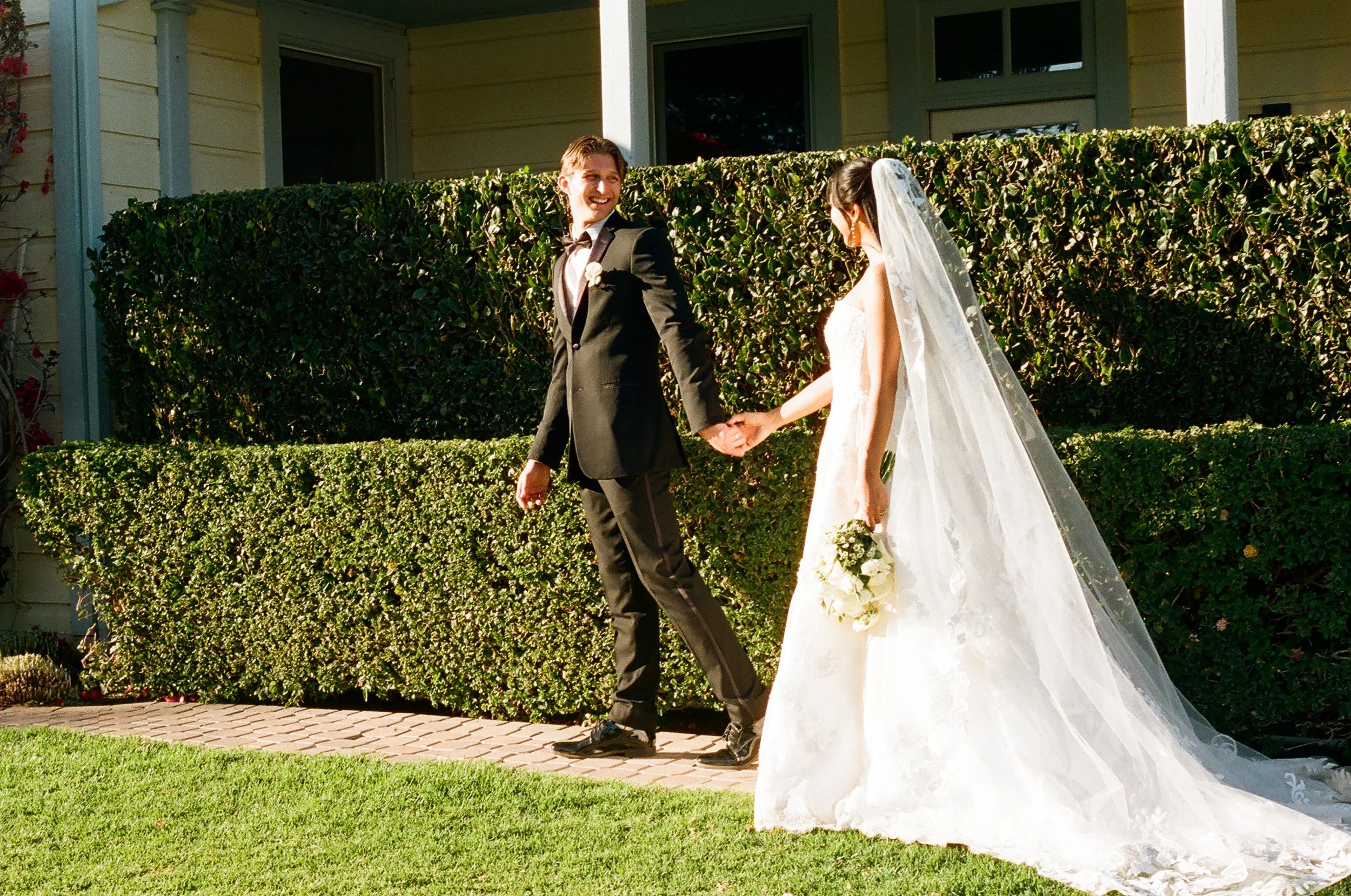 A smiling groom holding hands with a bride wearing a white wedding dress and veil, walking outside on a sunny day with green bushes and a house in the background.