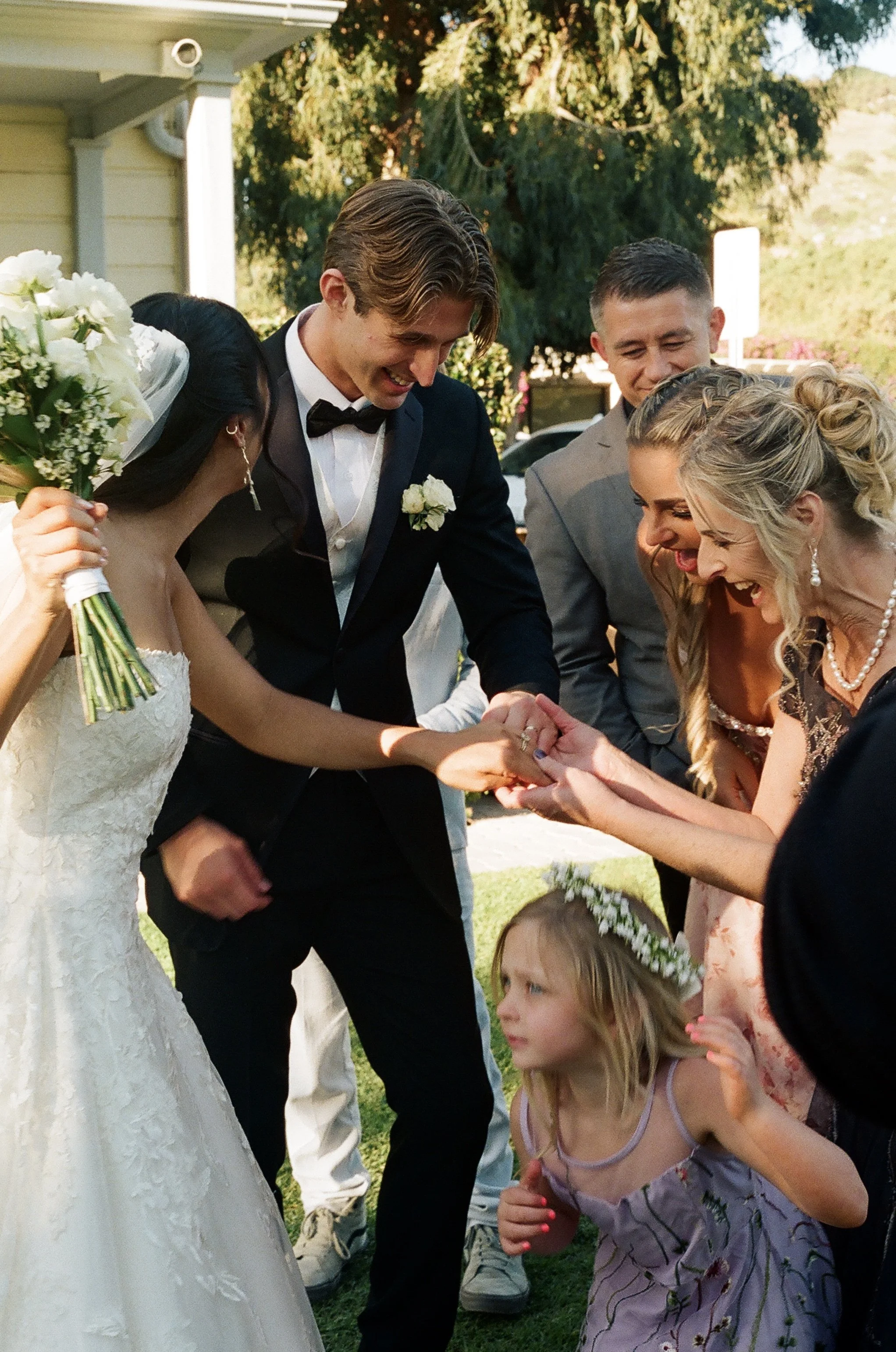 A wedding scene with a bride, groom, and guests outdoors. The bride holds a bouquet, the groom wears a tuxedo with a bow tie, and the guests are smiling and looking at the couple. Trees and a house are in the background.