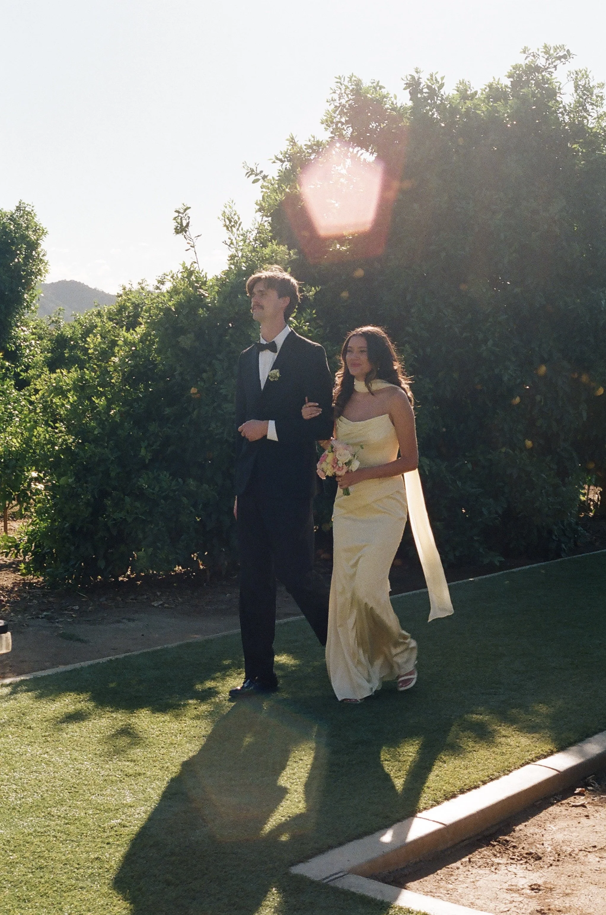 A man in a tuxedo and a woman in a yellow gown walking outdoors in a garden during daylight, with trees and a mountain in the background.