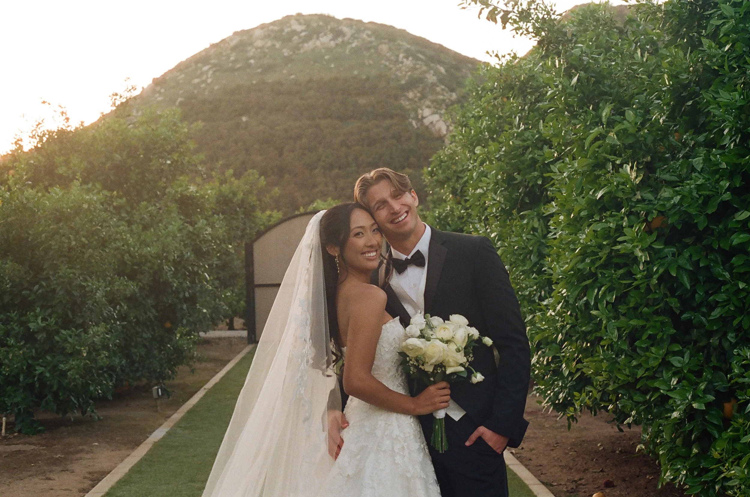 A bride and groom smiling and posing outdoors during their wedding, with greenery and mountain in the background at sunset.