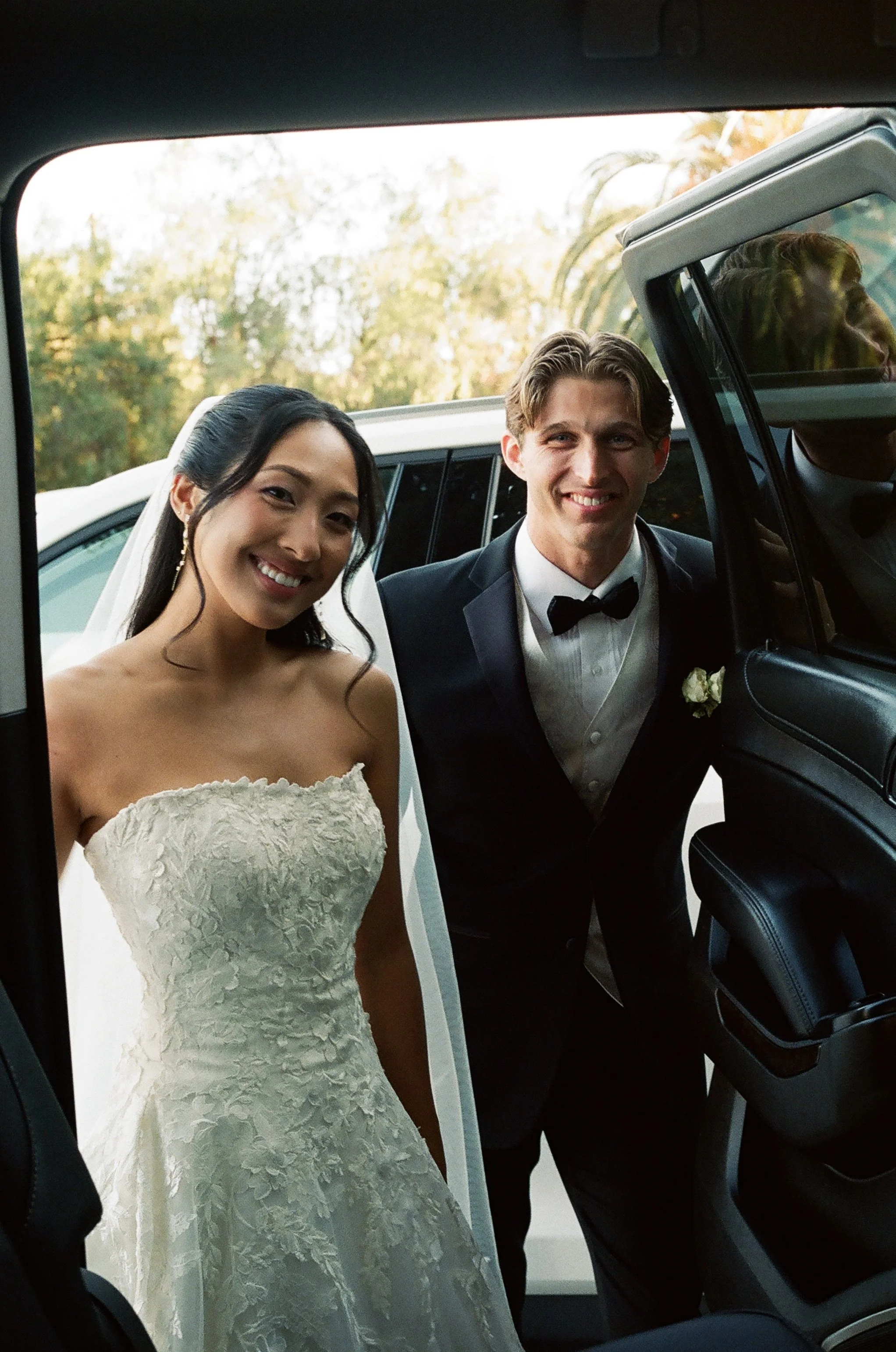 A bride and groom smiling and looking into the camera, standing outside their wedding vehicle, with a background of trees and a clear sky.