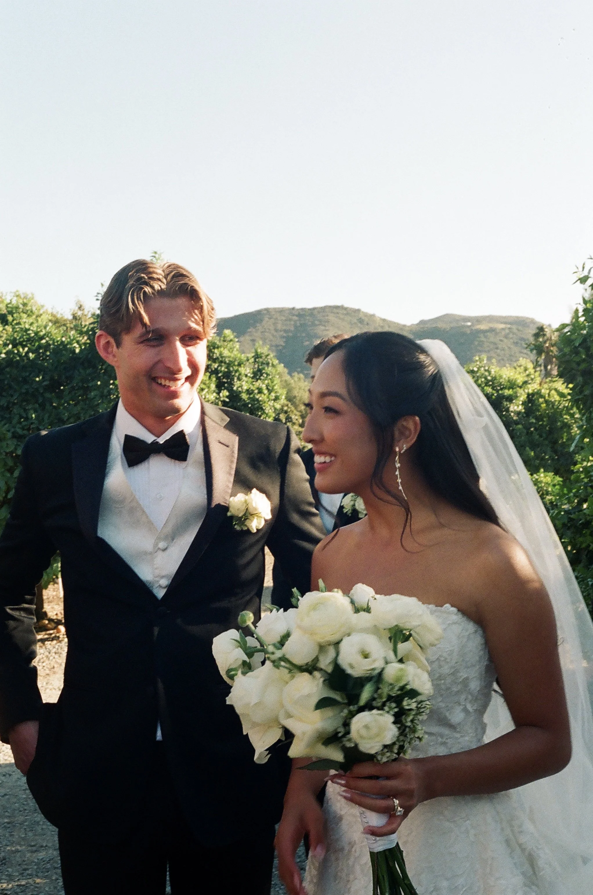 A smiling groom in a black tuxedo with a bow tie standing next to a smiling bride in a strapless wedding gown holding a bouquet of white flowers. They are outdoors with green bushes and hills in the background, under clear skies.