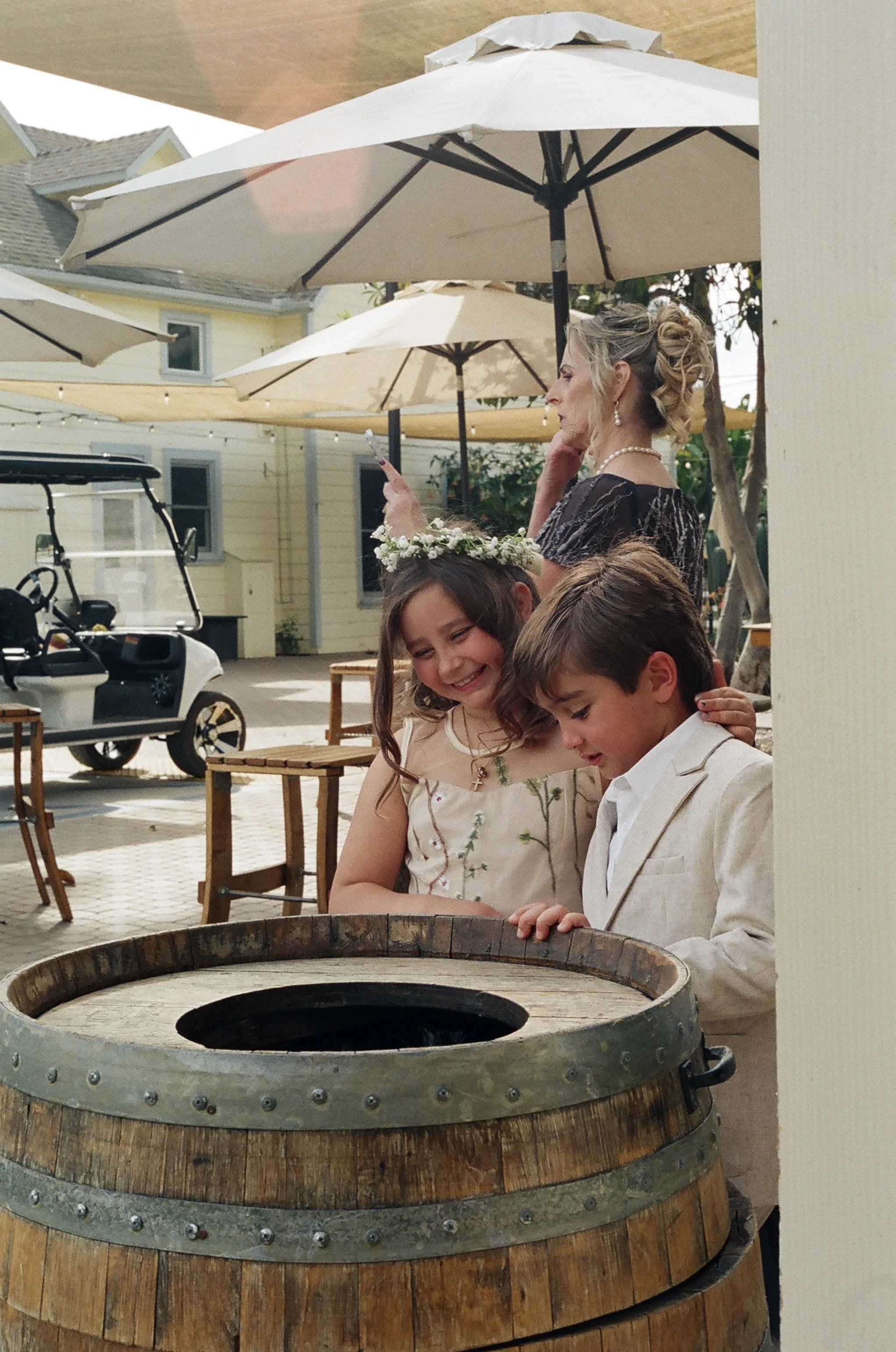 A woman and two children outside under umbrellas, with a large wooden barrel in the foreground, and a golf cart parked nearby, in a backyard setting during daytime.