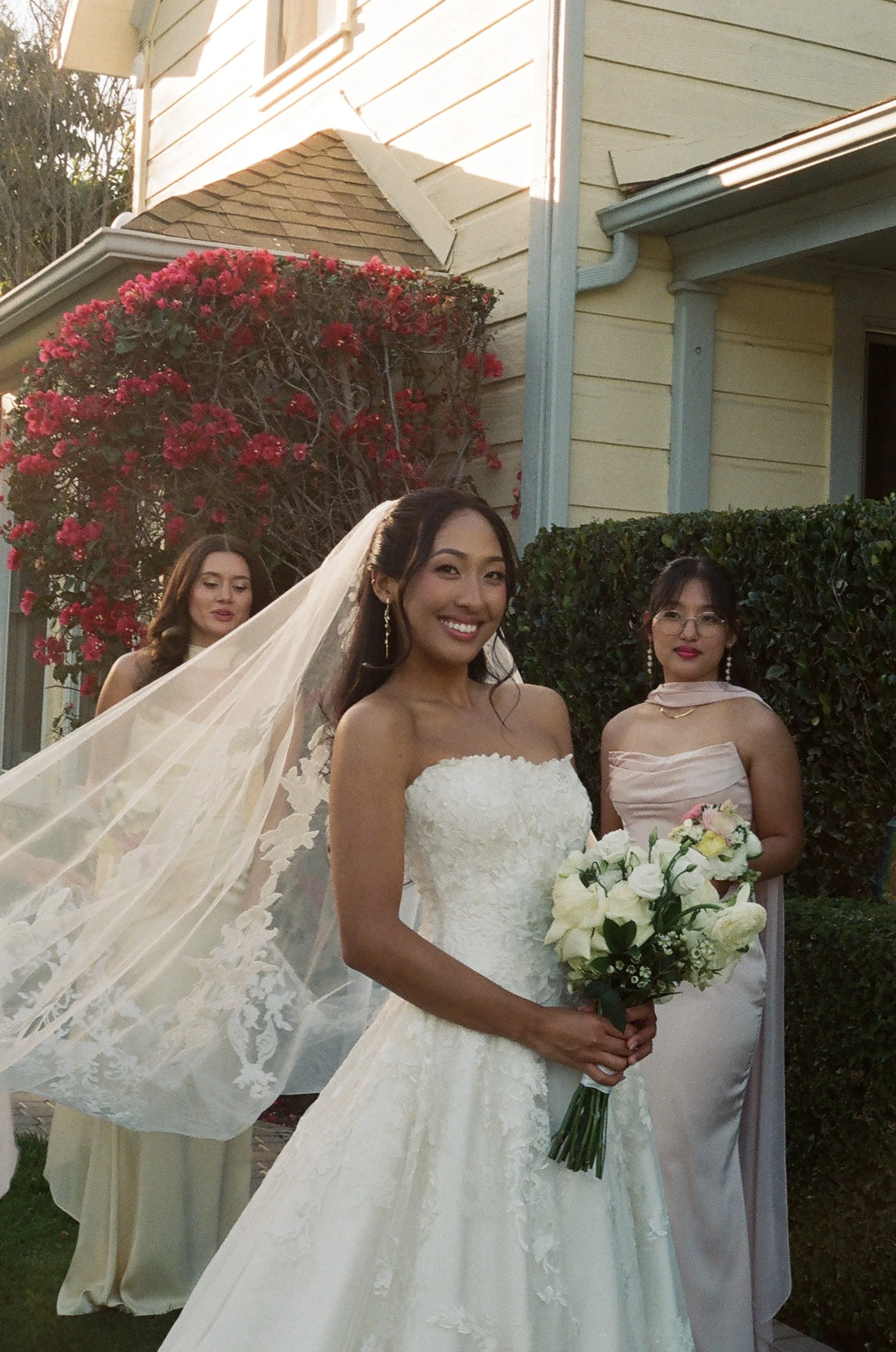 A bride in a white lace wedding dress holding a bouquet of white and yellow flowers, standing outdoors with a house and blooming red flowers in the background. She is surrounded by women in formal dresses at a wedding ceremony.
