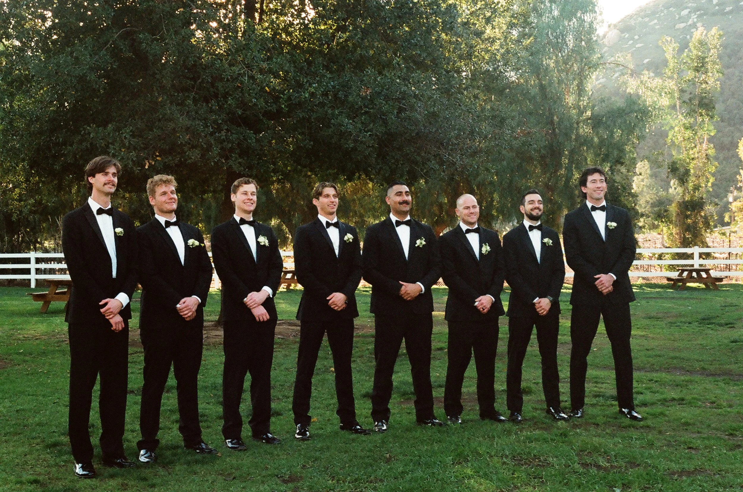 Line of nine men dressed in black tuxedos with white shirts and black bow ties, standing outdoors on a grassy area with trees in the background, during daytime.