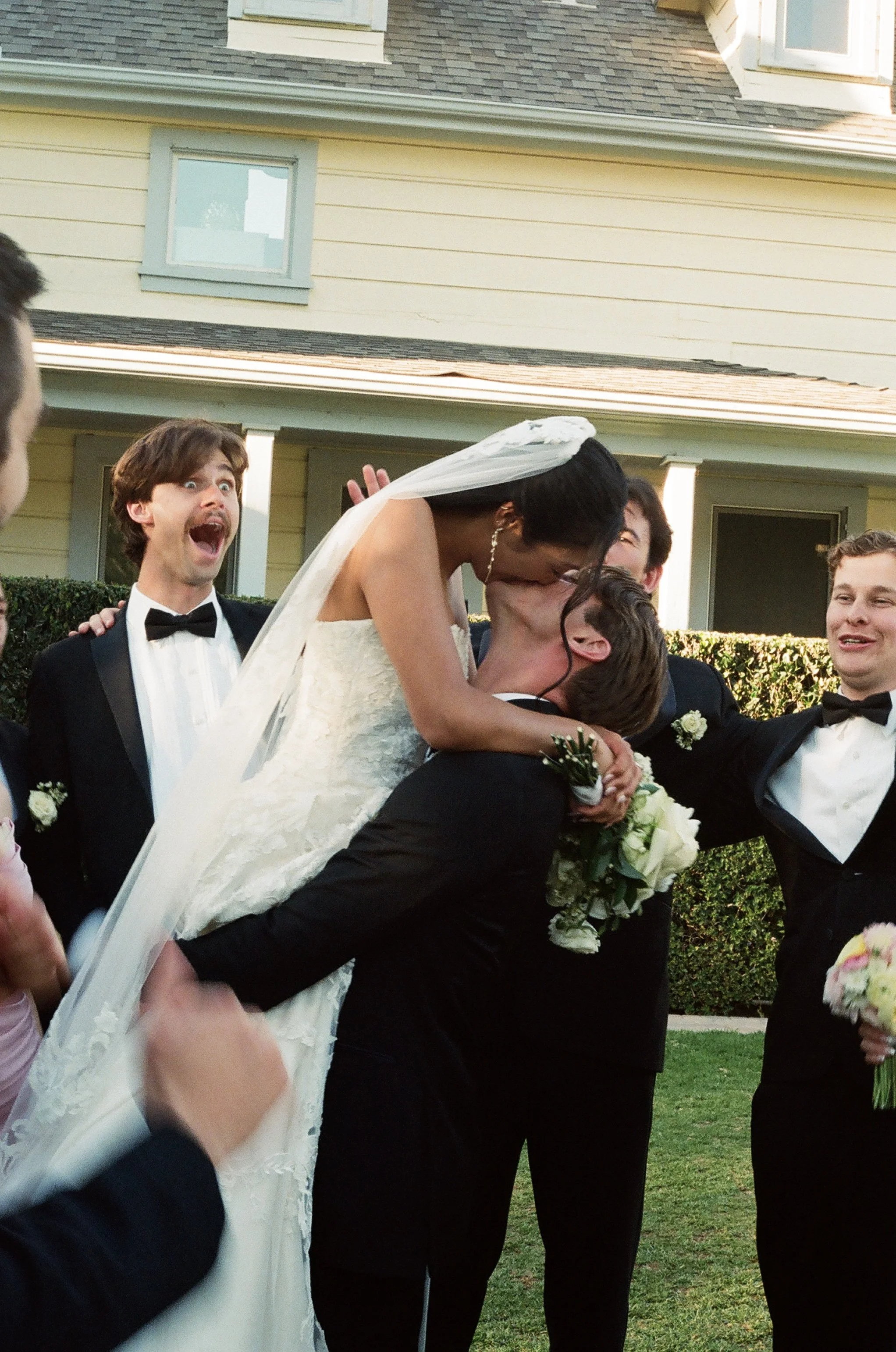 A newlywed couple shares a kiss while being lifted by friends during an outdoor wedding celebration.