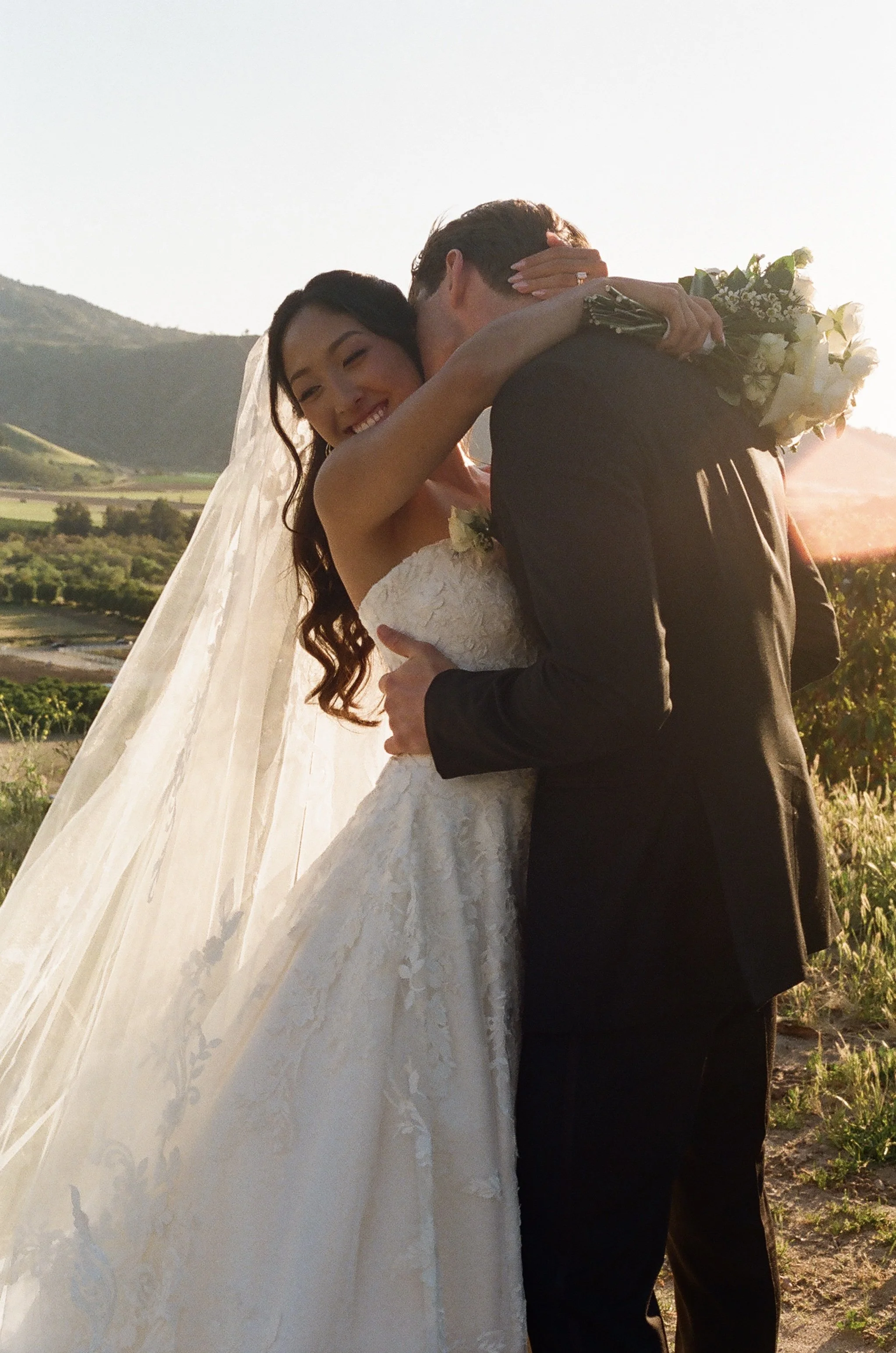 A bride and groom embrace outdoors during sunset, with the bride smiling and holding a bouquet, in a scenic rural landscape with hills and fields in the background.