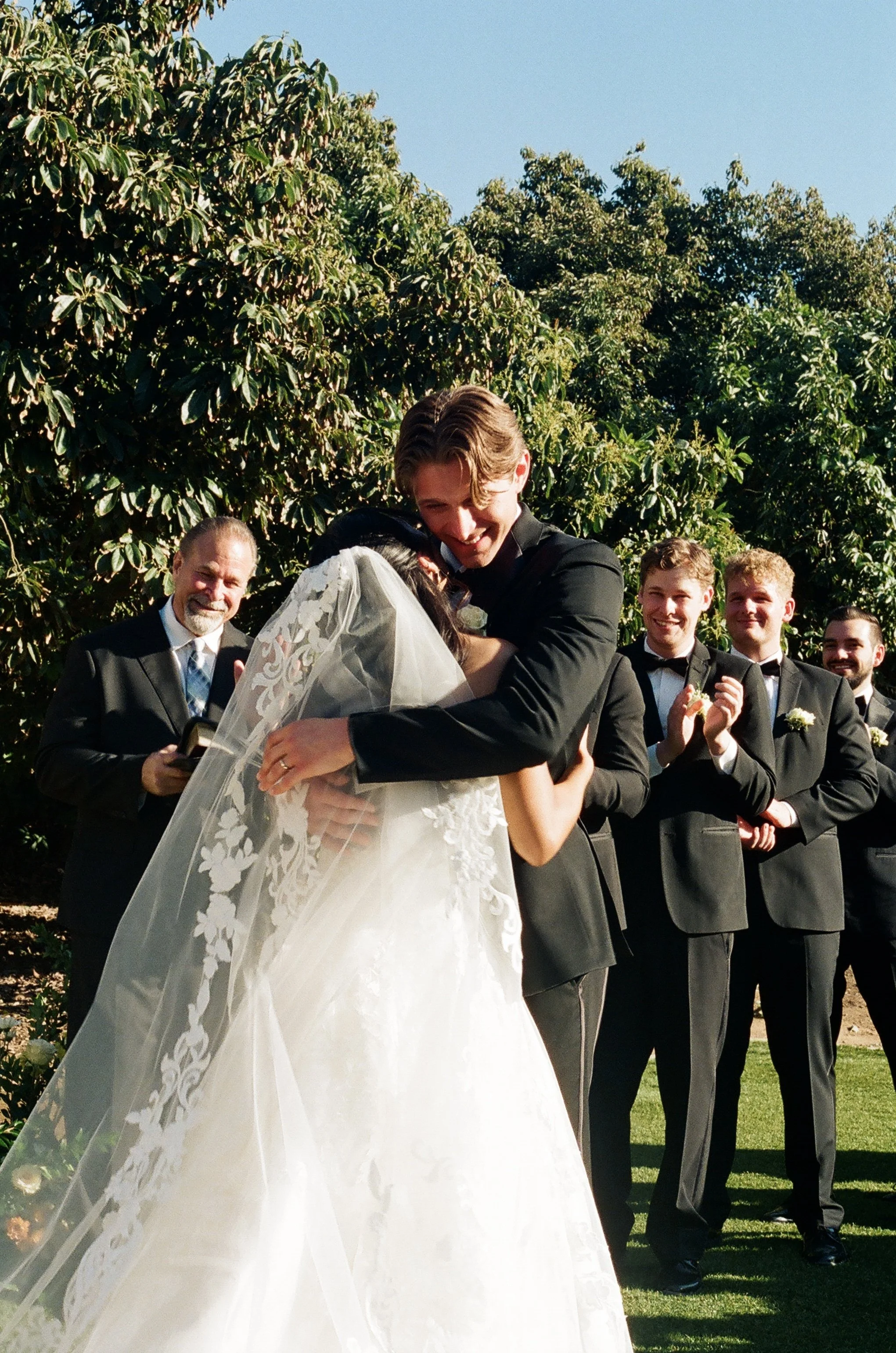 A wedding ceremony outdoors with a bride and groom embracing, surrounded by groomsmen clapping, on a sunny day with trees in the background.