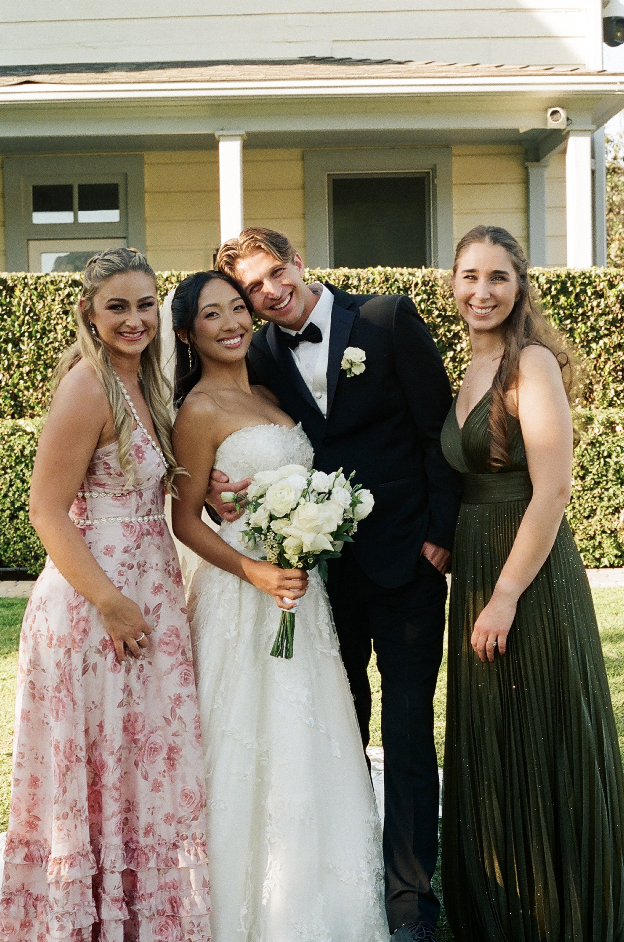 Group of four people, two women and two men, standing outdoors in front of a house with a hedge behind them. The woman in the center is wearing a white wedding dress and holding a bouquet of white roses. The man next to her is dressed in a black tuxe