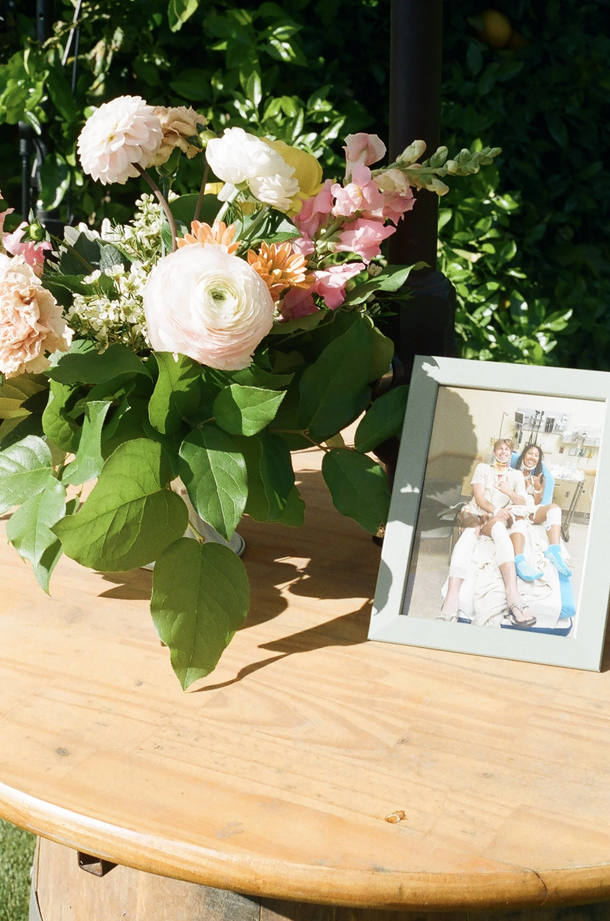 A bouquet of various flowers, including pink, white, and orange blooms, placed on a wooden surface next to a framed photo of two women smiling and sitting together, outdoors with green foliage in the background.
