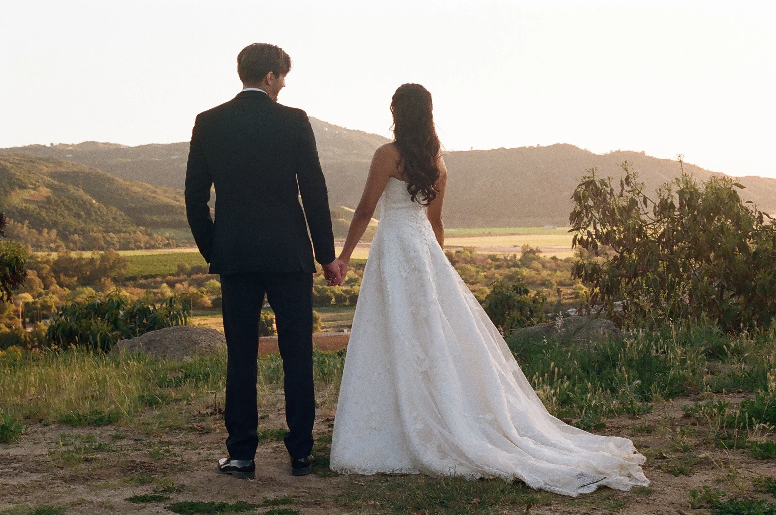 A bride and groom holding hands outdoors with mountains in the background, during sunset.