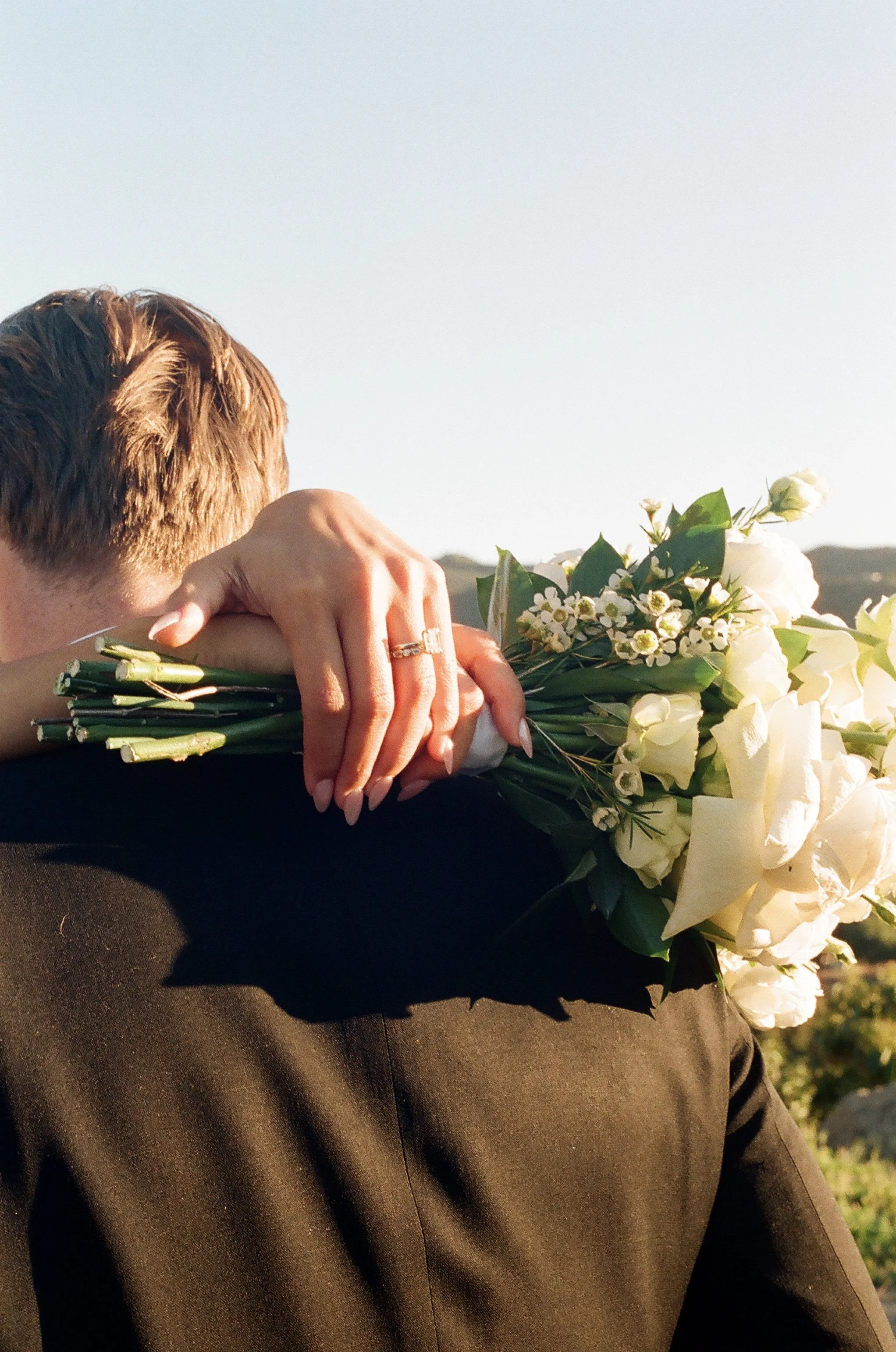 A person with short brown hair holding a bouquet of white flowers, wearing a black outfit, outdoors during sunset or sunrise.