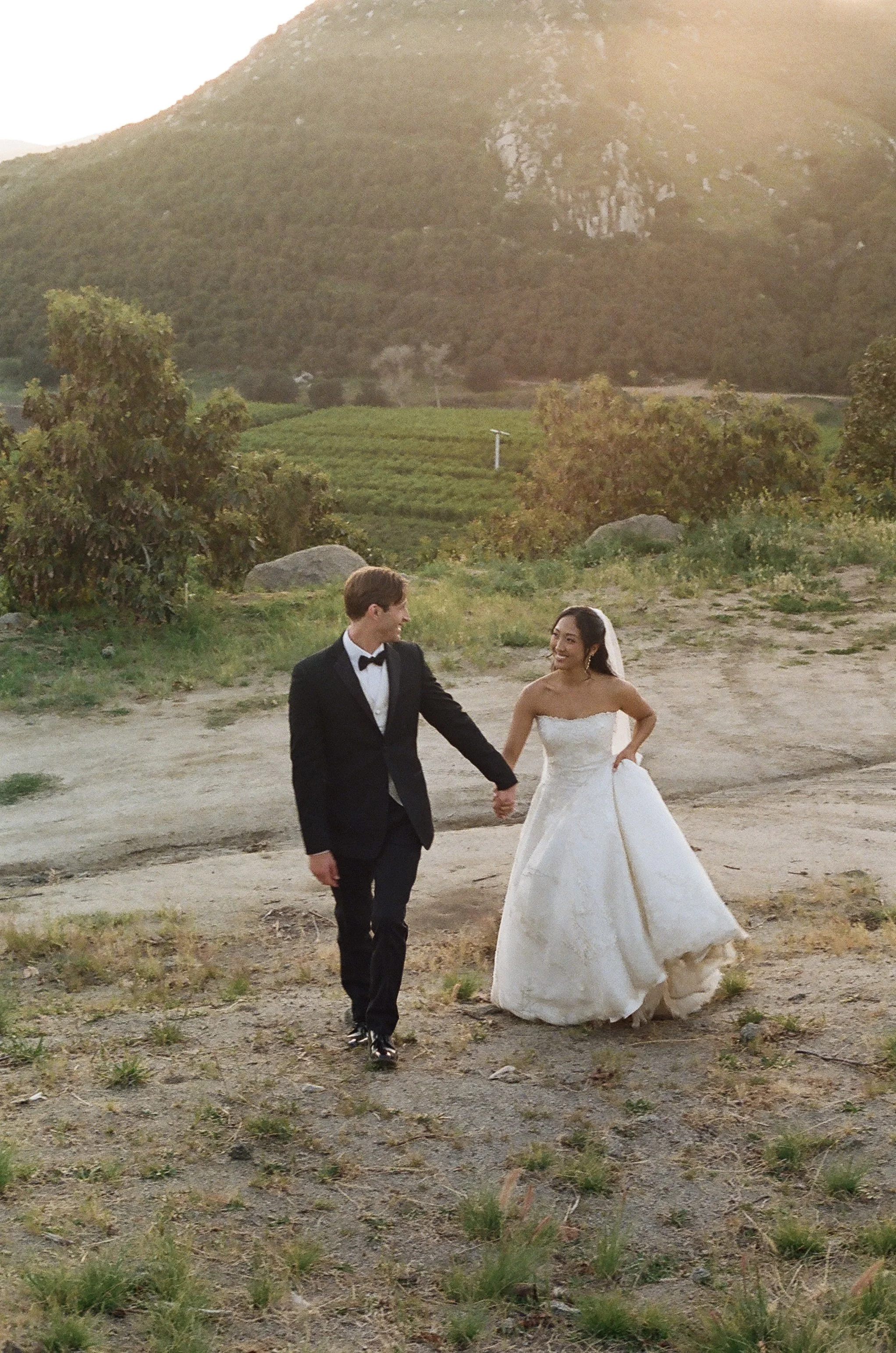 A bride and groom walking hand in hand outdoors near a mountain, with trees and greenery in the background, during a sunset.