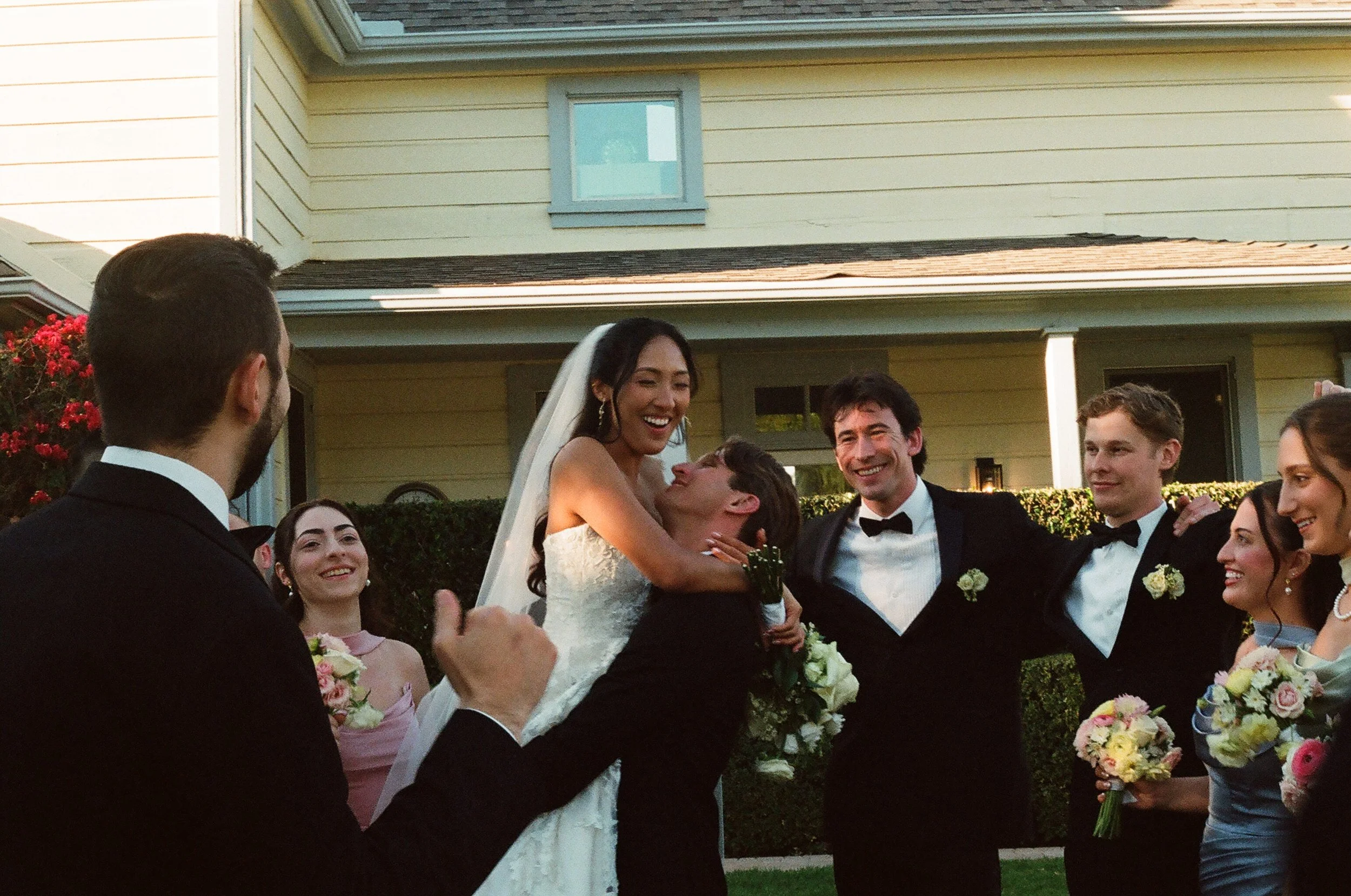 A group of wedding guests celebrating outdoors in front of a yellow house with green bushes. The bride, wearing a white wedding dress and veil, is smiling and being lifted by a man in a tuxedo. Other guests are holding bouquets and smiling.