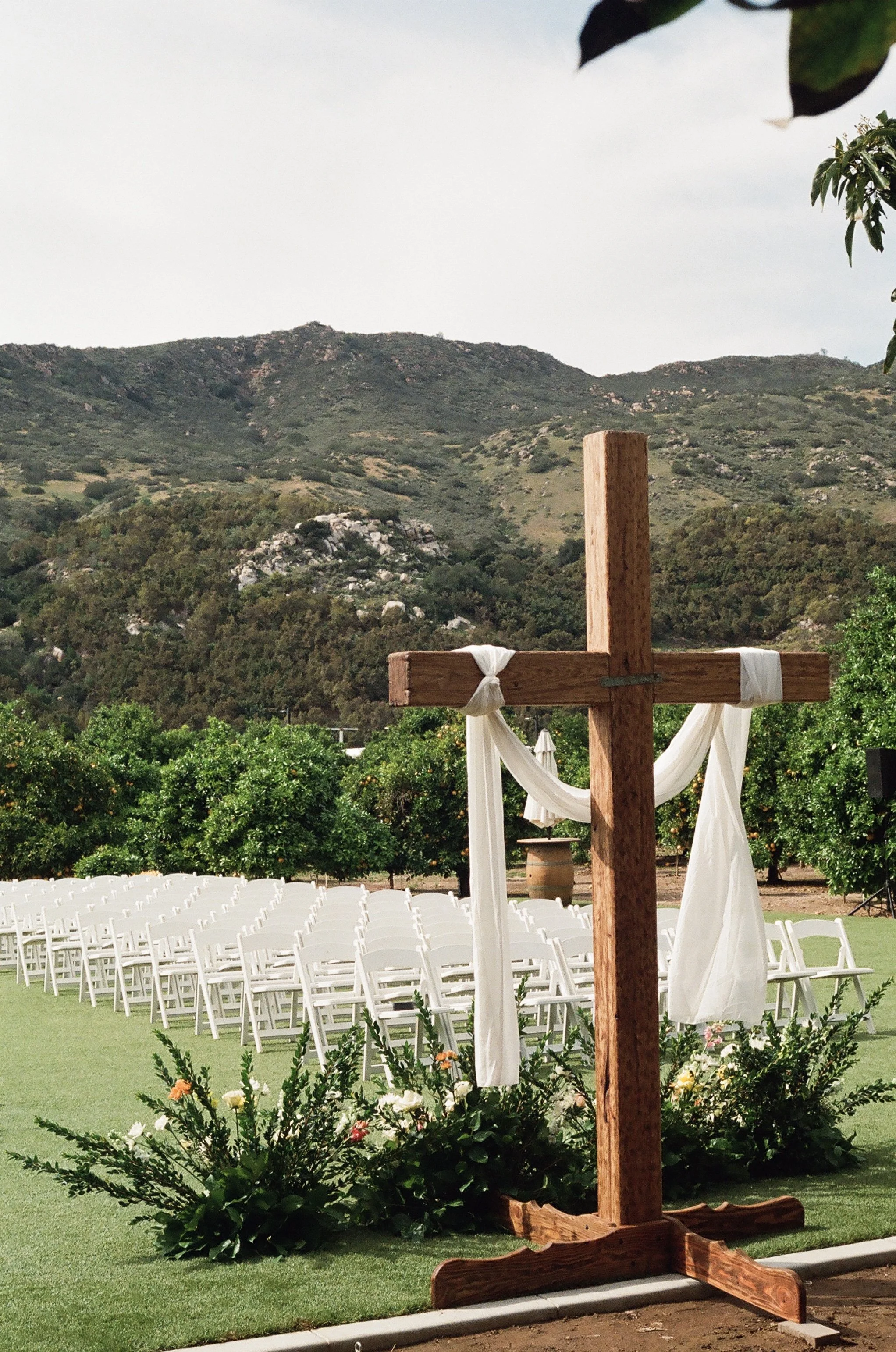 Outdoor wedding ceremony setup with rows of white chairs and a wooden cross decorated with white fabric and flowers, set against a backdrop of mountains and trees.