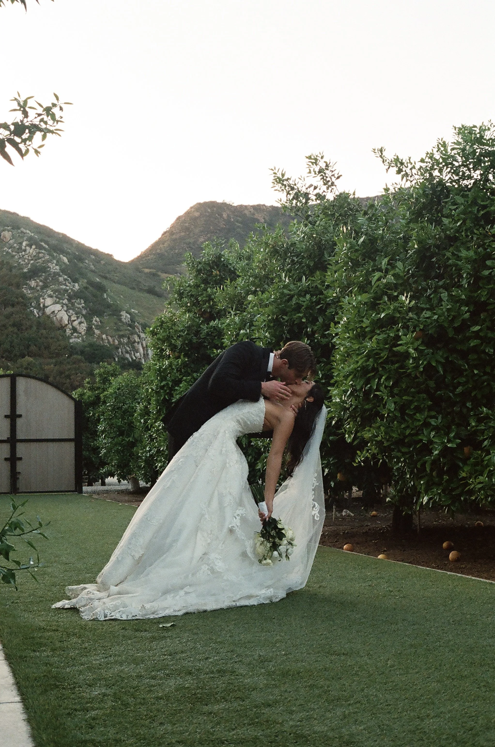 A newlywed couple sharing a kiss outdoors with a background of mountains, green trees, and a partly cloudy sky, with the bride holding a bouquet of white flowers.