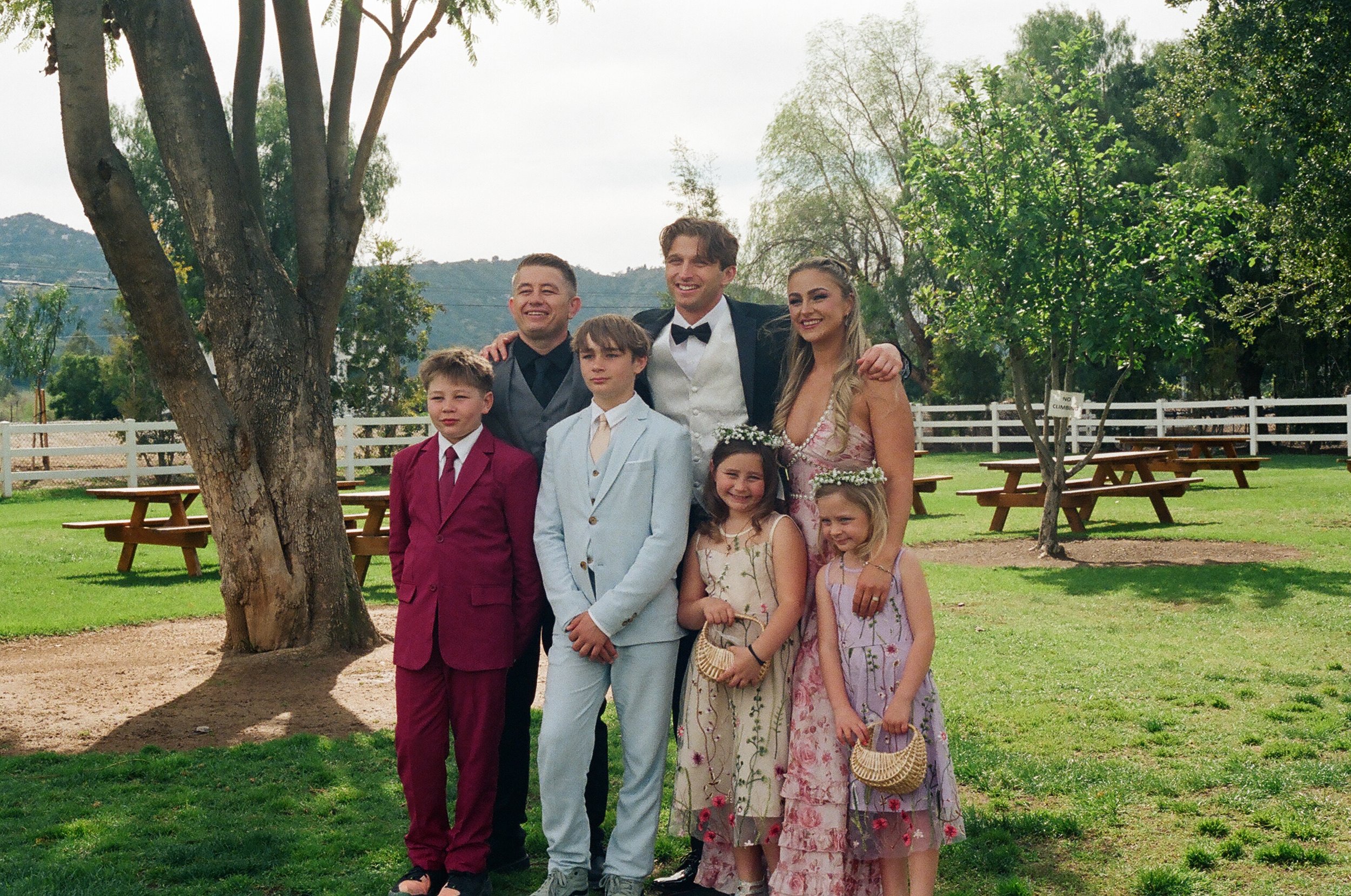 Group of seven people, including children and adults, standing outdoors in a park-like setting with picnic tables, trees, and mountains in the background. Some adults are wearing formal attire, indicating a celebration or special occasion.
