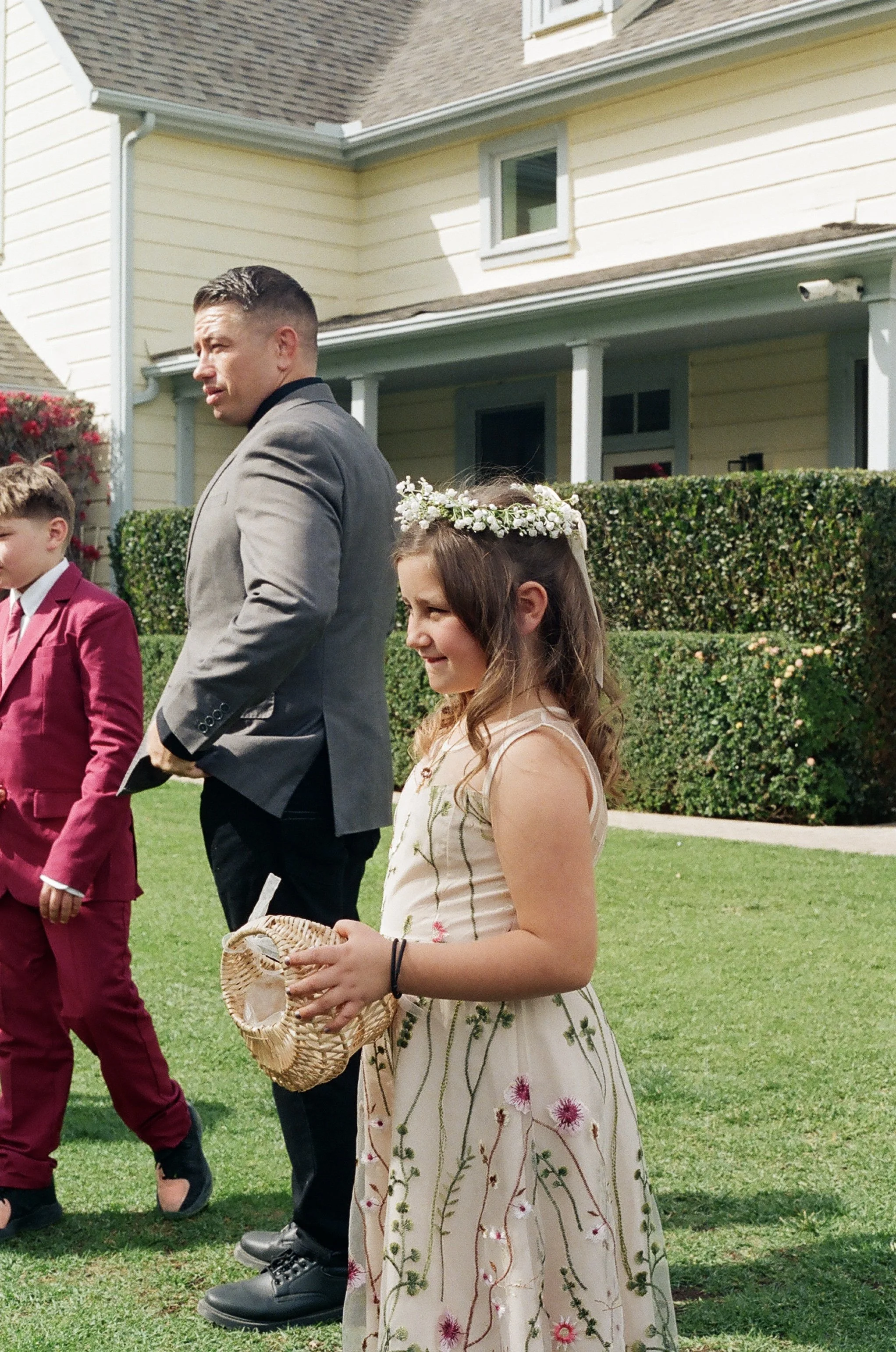 Young girl with long hair, floral dress, and floral headband holding a wicker basket, standing outdoors in front of a yellow house with manicured bushes. Part of a man in a gray suit and young boy in a pink suit are visible beside her.