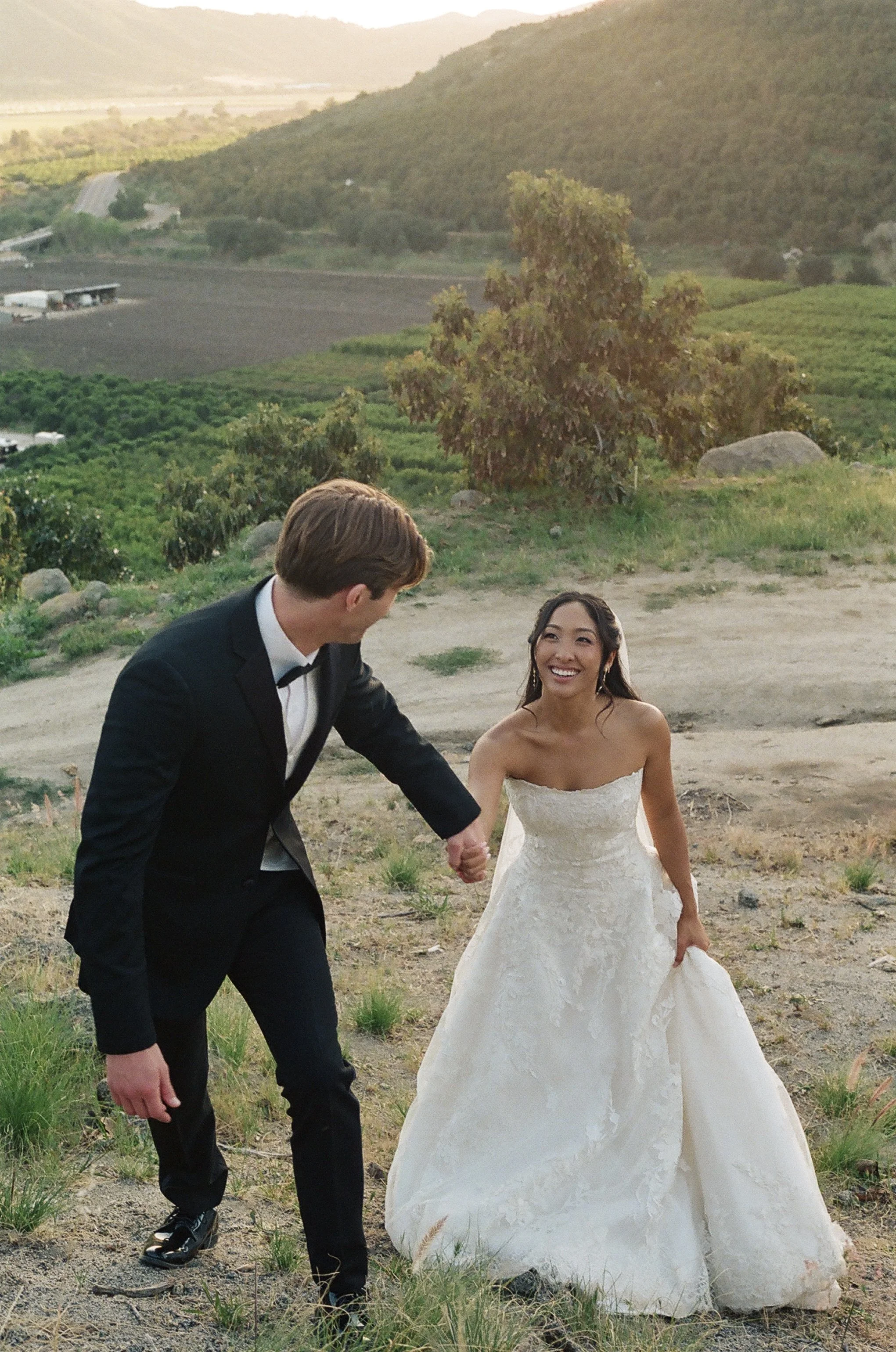 A man in a tuxedo and a woman in a white wedding dress holding hands outdoors, with a scenic landscape of rolling hills, farmland, and distant mountains during sunset.