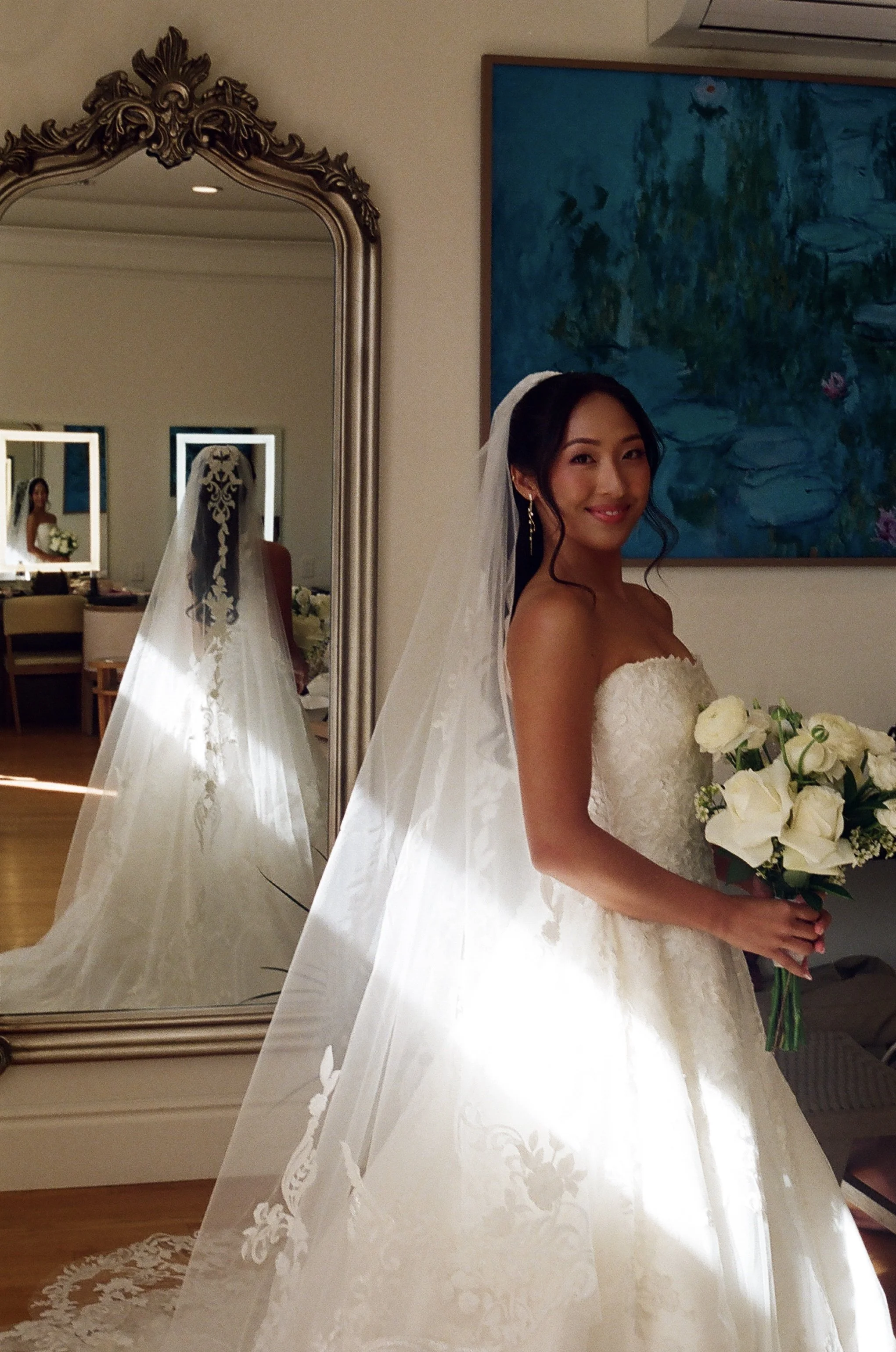 Brunette bride in a white wedding gown holding a bouquet of white roses, standing near a large mirror with ornate frame, with a colorful painting on the wall behind her.