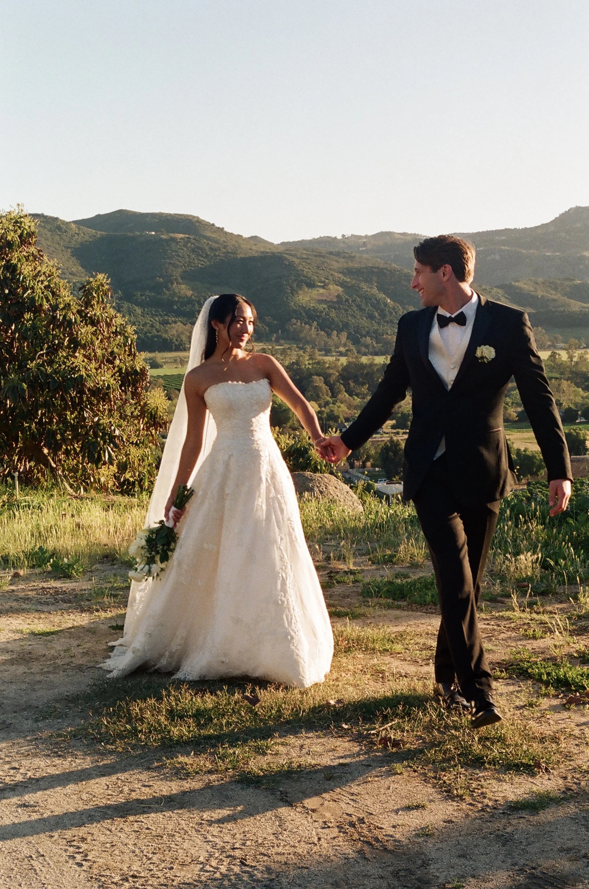 A bride and groom holding hands outdoors in a scenic, hilly landscape during sunset, dressed in wedding attire, smiling at each other.