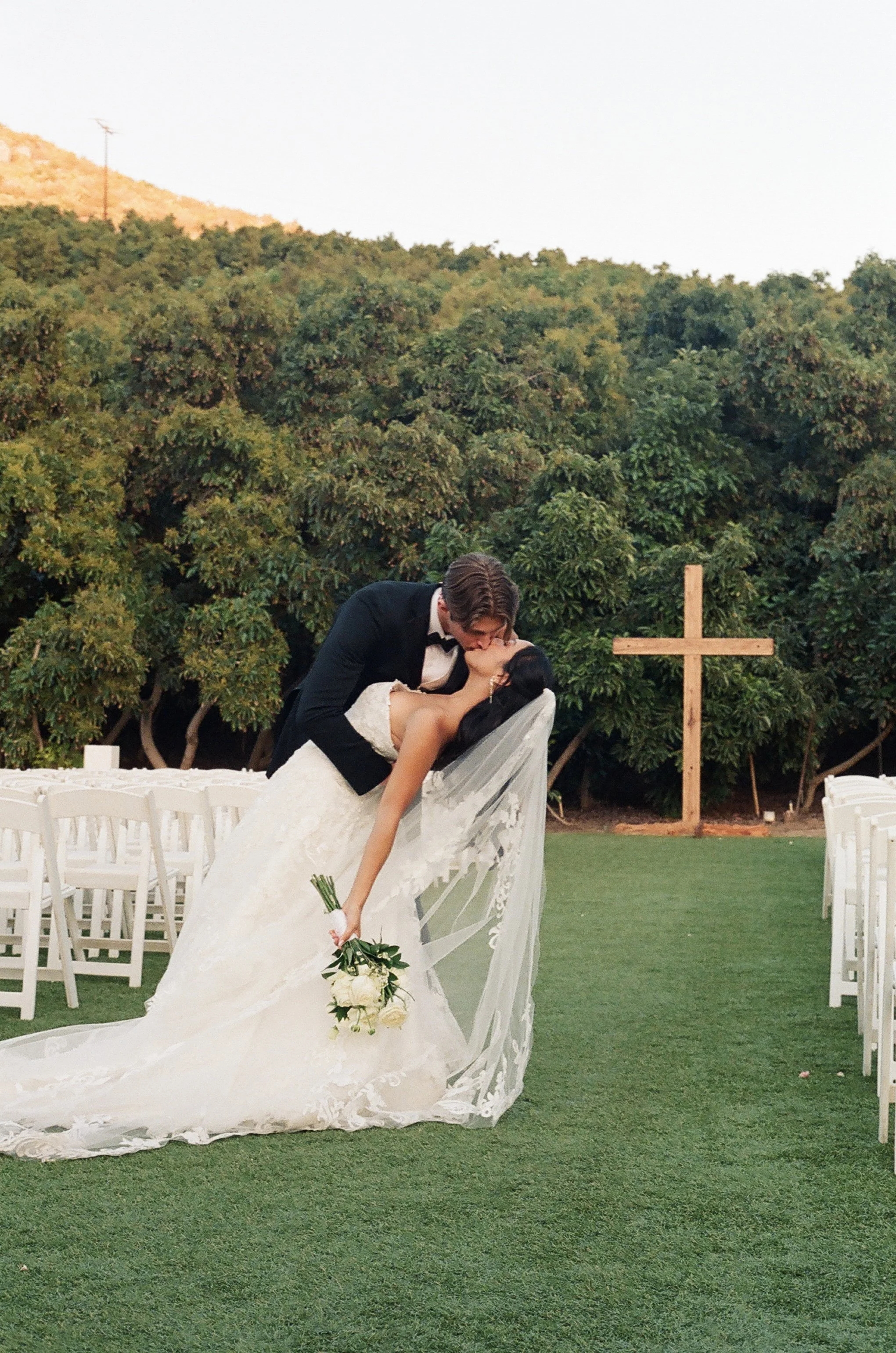 A bride and groom share a kiss during their outdoor wedding ceremony, with the bride holding a bouquet of white flowers and the groom wearing a black tuxedo. Green trees and a cross are visible in the background.