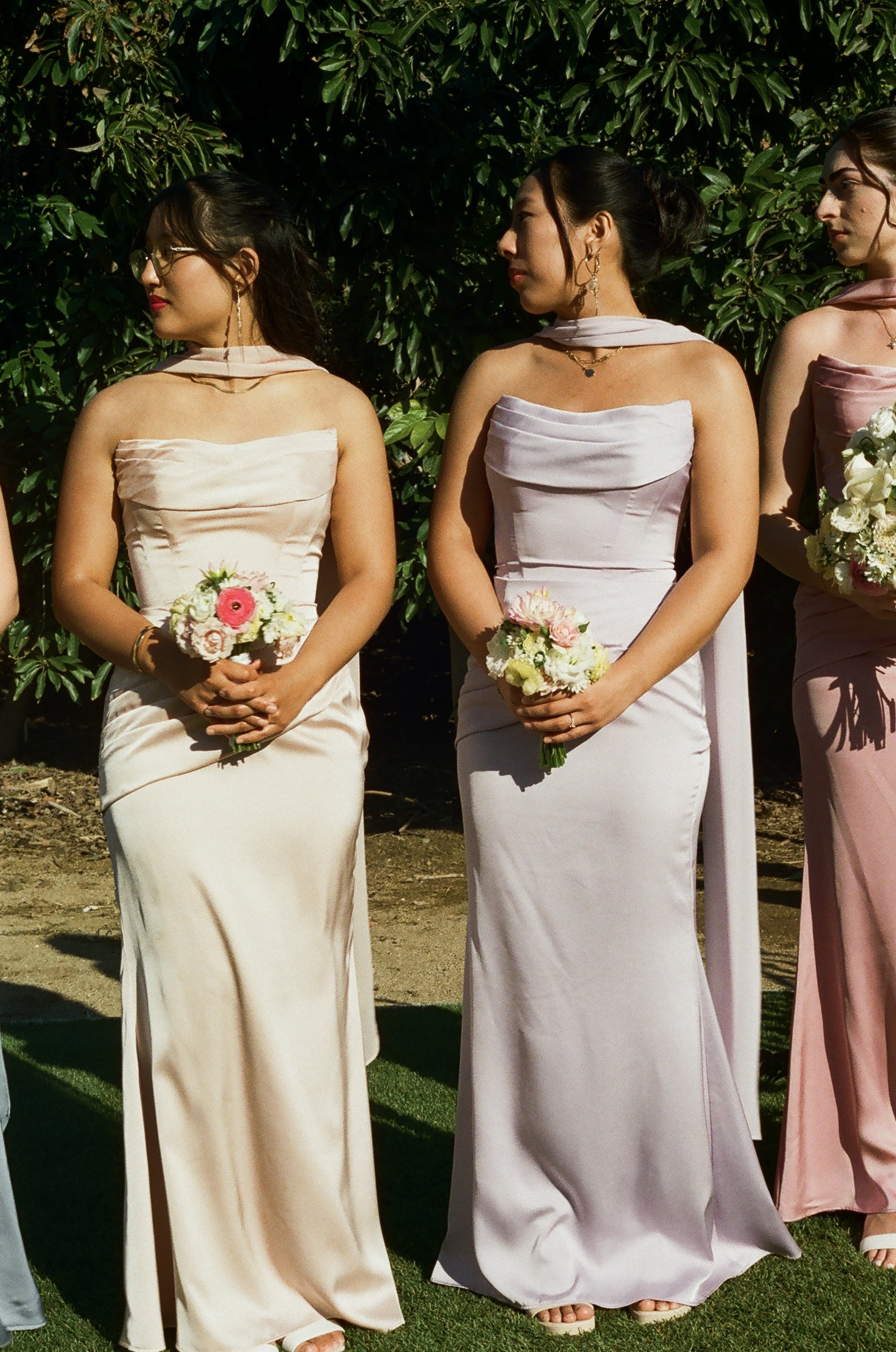 Women in pastel-colored dresses holding bouquets, standing outdoors near greenery.