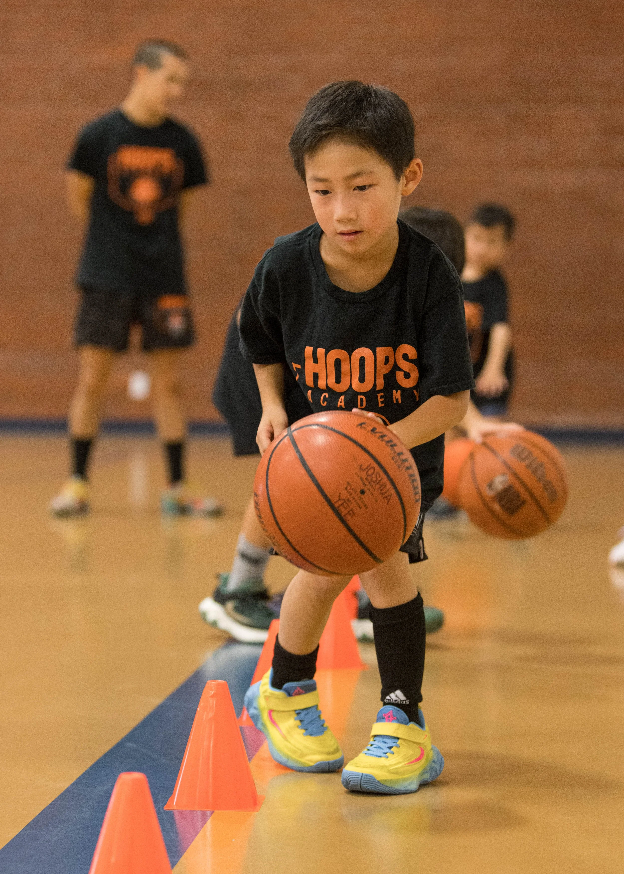 Young boy in a blue basketball uniform with a number 8, holding a basketball in a gymnasium with players in the background.