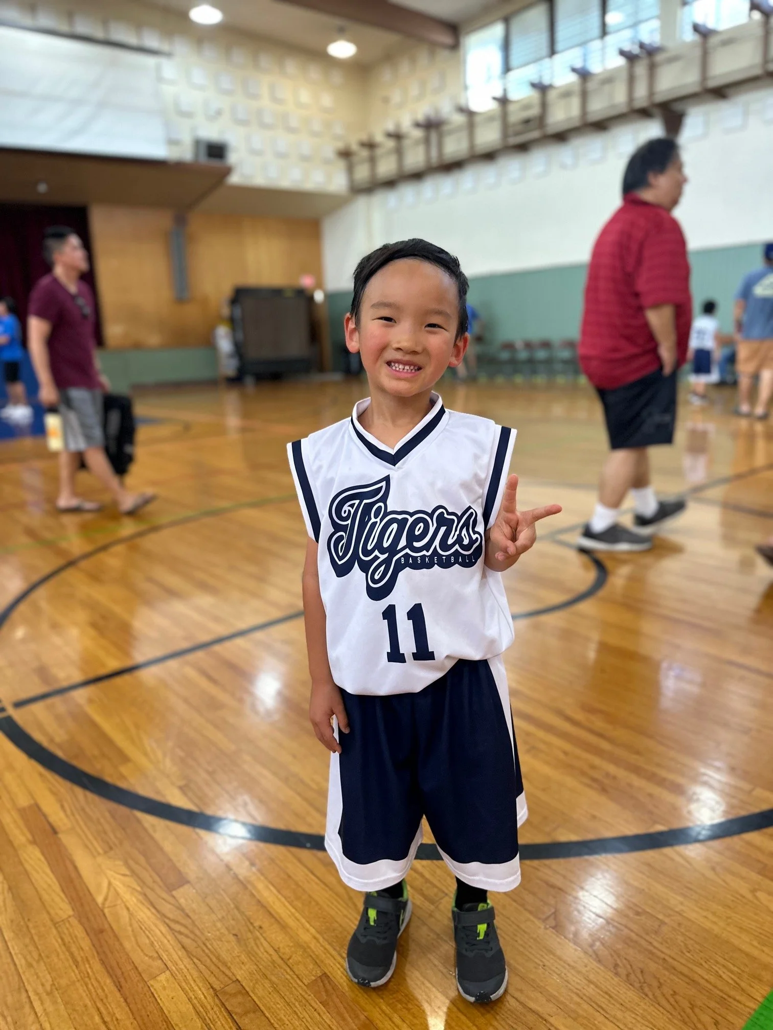 A young boy in a white and blue basketball uniform with the number 11, smiling and standing on a gymnasium basketball court.
